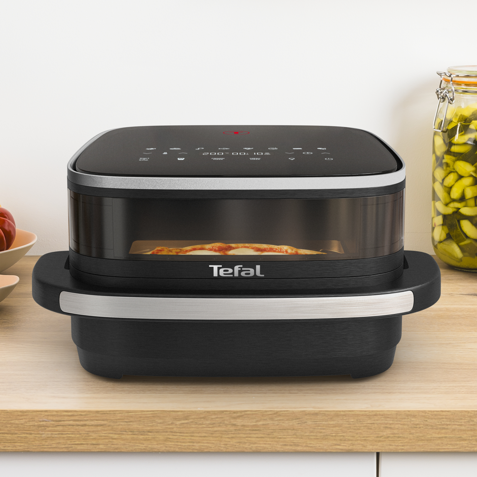 Czarny i srebrny piekarnik z pizzą w środku, ustawiony na drewnianej powierzchni z innymi przedmiotami.