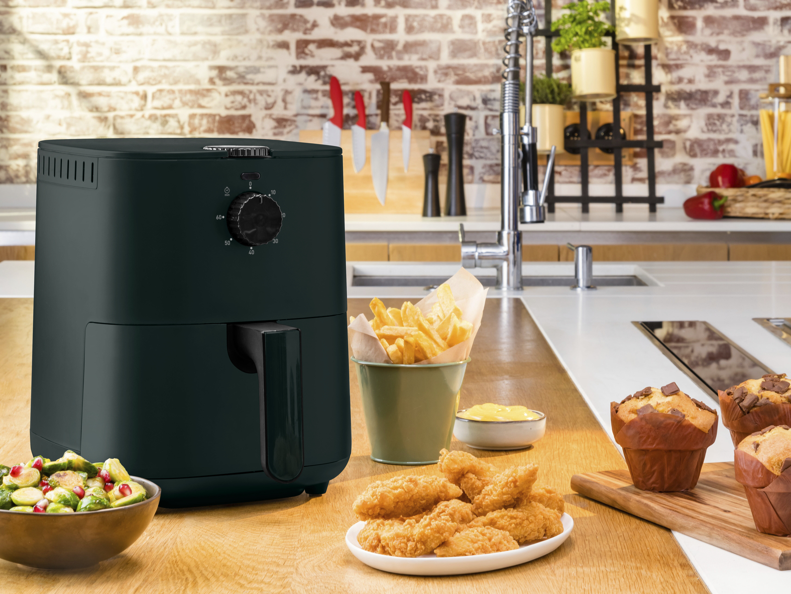 Una friggitrice ad aria con cibi fritti e muffin su una superficie di legno, cucina sullo sfondo.