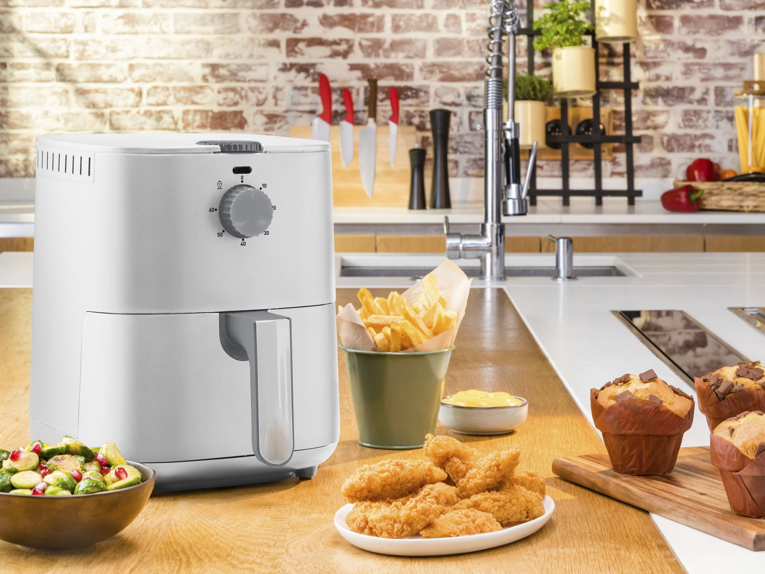 Una friggitrice ad aria con patatine fritte, pepite di pollo, muffin e cavoletti di Bruxelles su un piano cucina in legno.