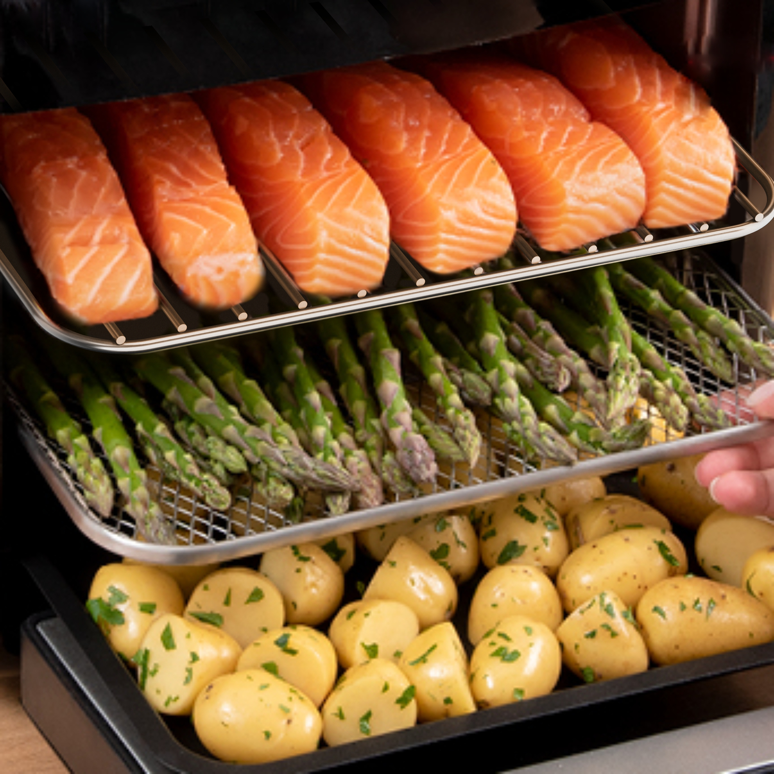 Frytkownica powietrzna z warstwami jedzenia: łosoś, szparagi i ziemniaki. Jedzenie jest ułożone na metalowych rusztach.