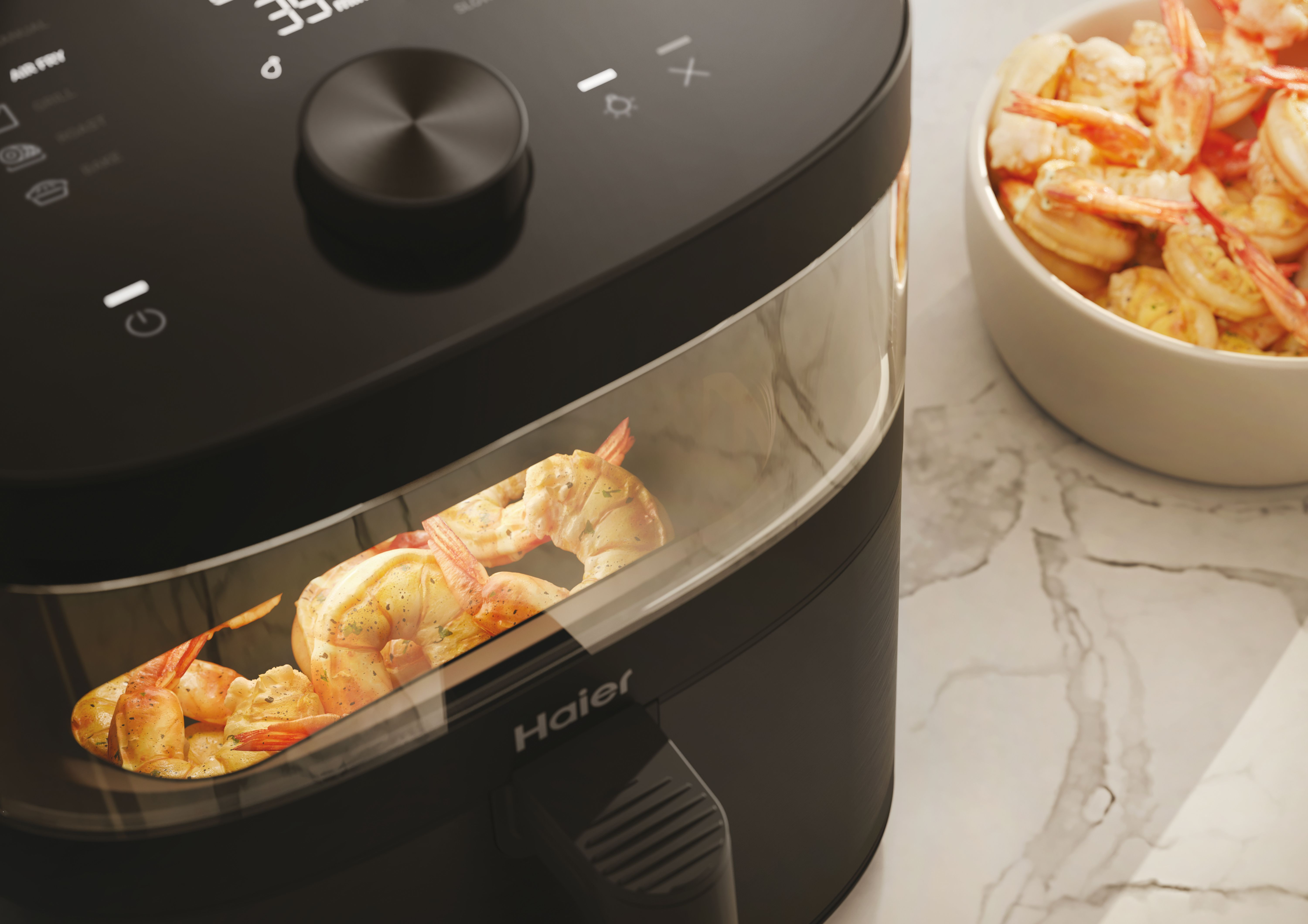 Friggitrice ad aria nera con gamberetti all'interno. Una ciotola di gamberetti cotti di lato. Logo Haier visibile.