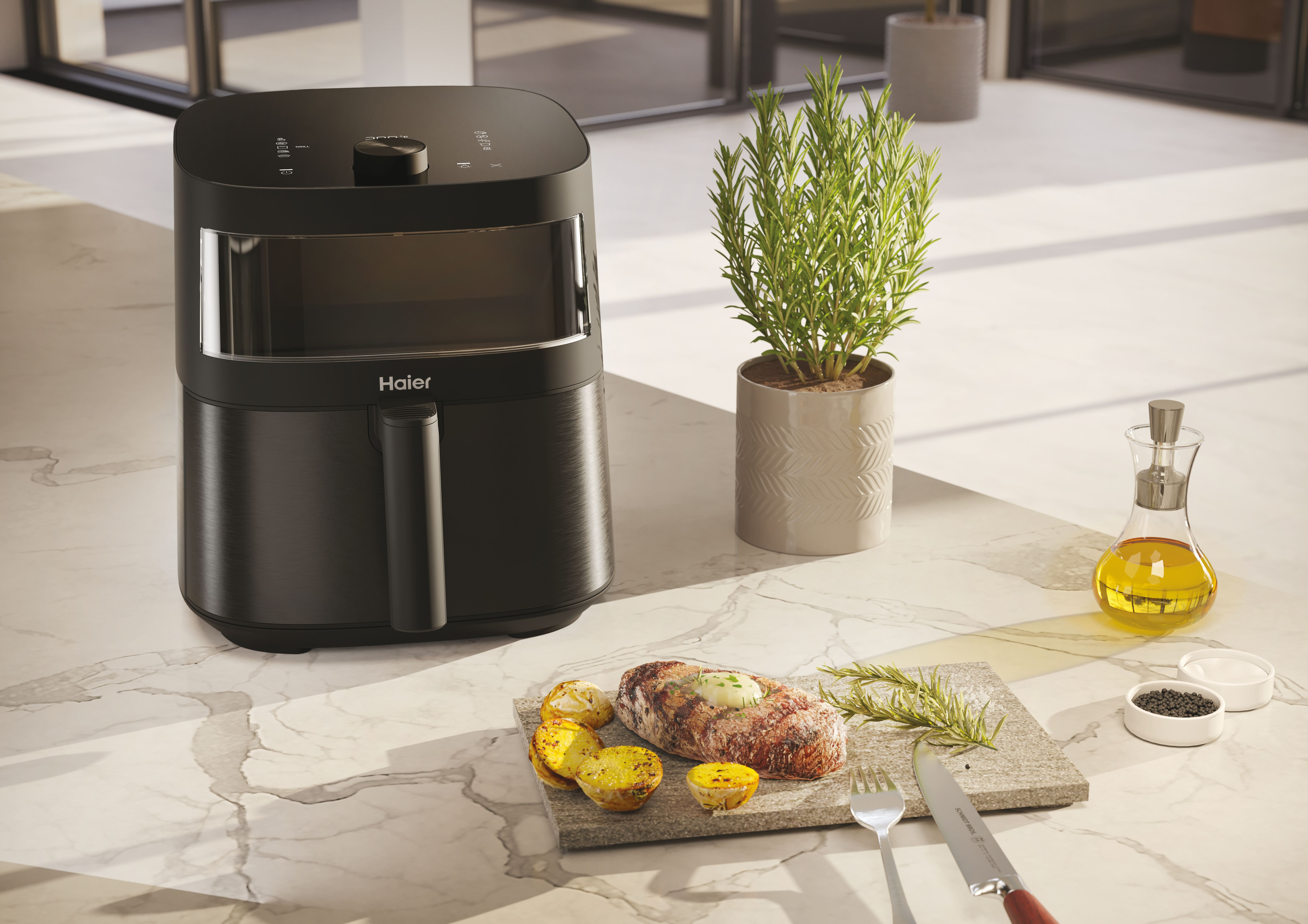 Una friggitrice ad aria nera con cibo e accessori su un bancone di marmo. Pianta, bistecca e bottiglia d'olio.