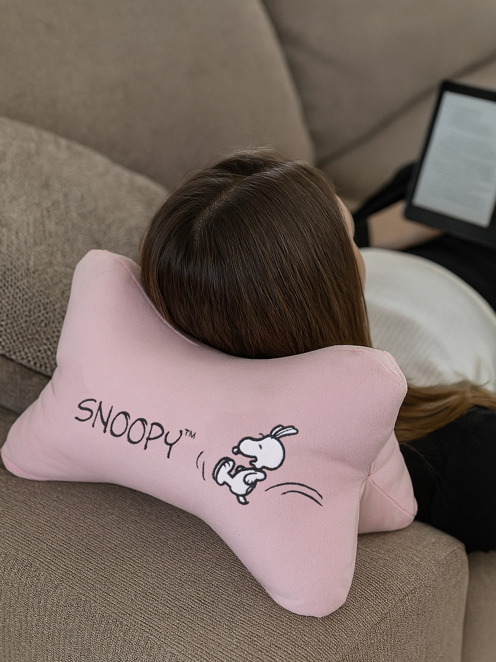 Eine Frau ruht auf einem Sofa mit einem rosa Snoopy-Kissen. Ein E-Reader ist im Hintergrund sichtbar.