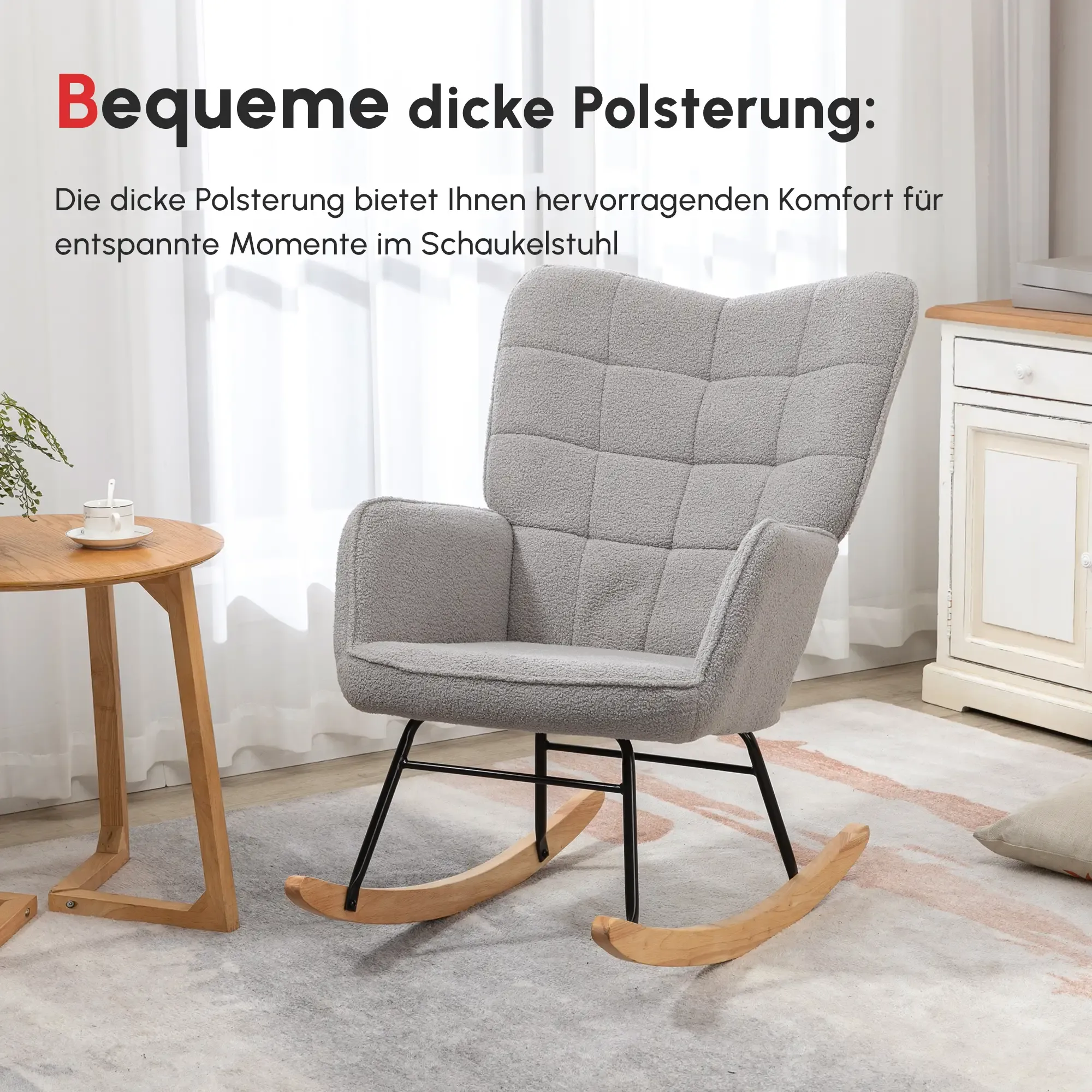 Ein grauer Schaukelstuhl steht auf einem Teppich. Ein runder Holztisch mit einer Tasse und einer Pflanze steht links.
