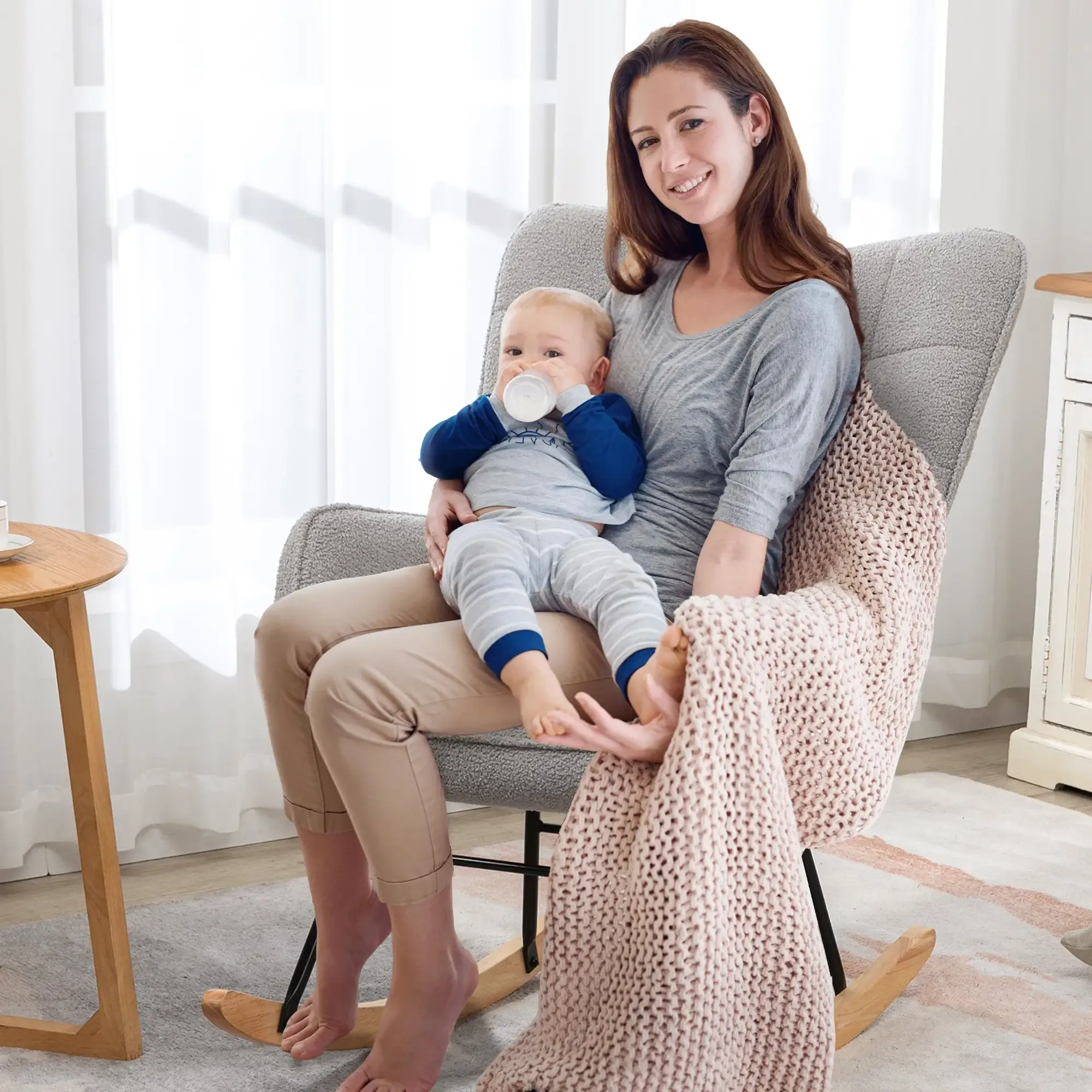 Eine Frau hält ein Baby, das mit einer Flasche gefüttert wird, in einem Schaukelstuhl sitzend. Eine Decke liegt auf dem Stuhl.