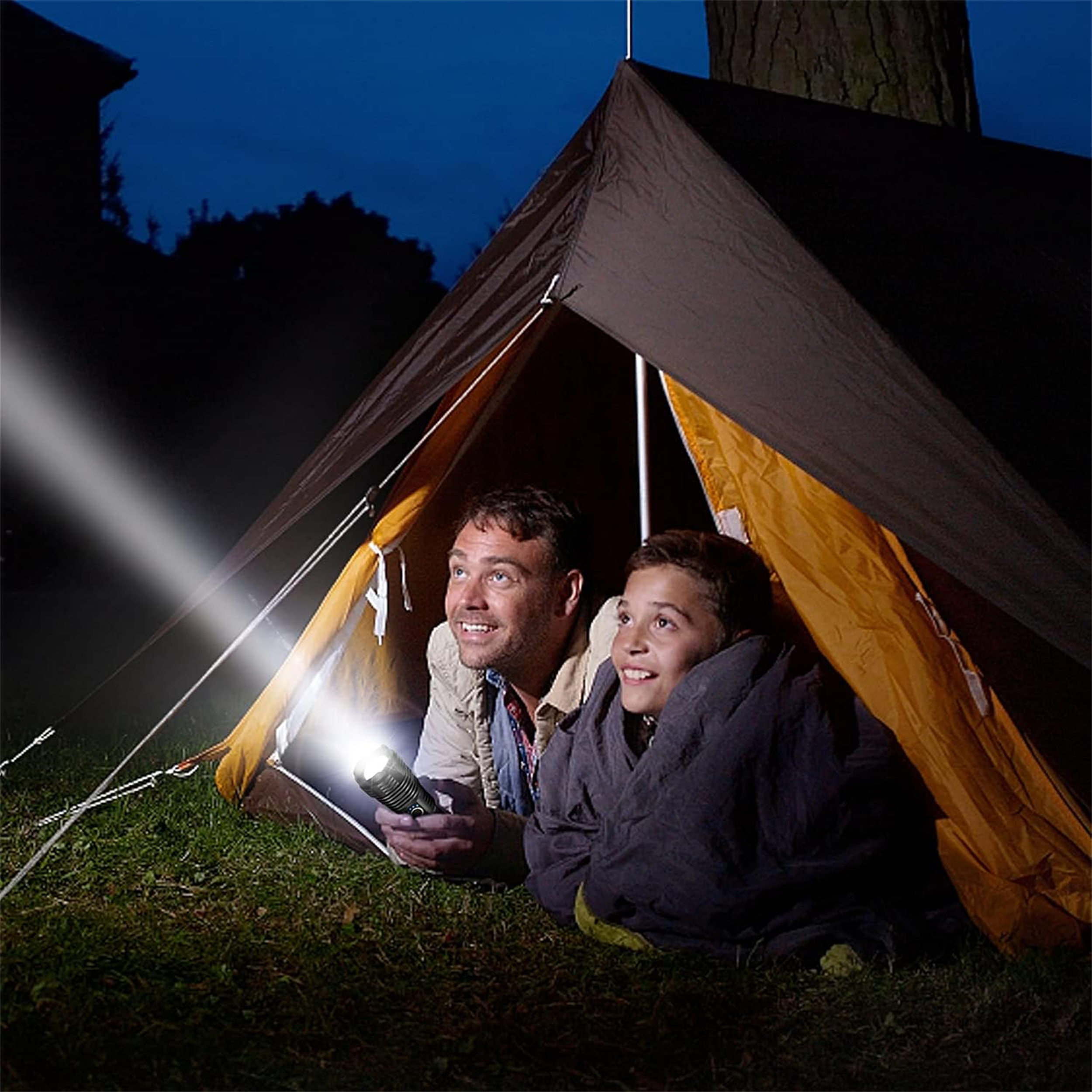 Zwei Personen in einem Zelt bei Nacht, beleuchtet von einer Taschenlampe. Dunkelblauer Himmel und grünes Gras sind sichtbar.