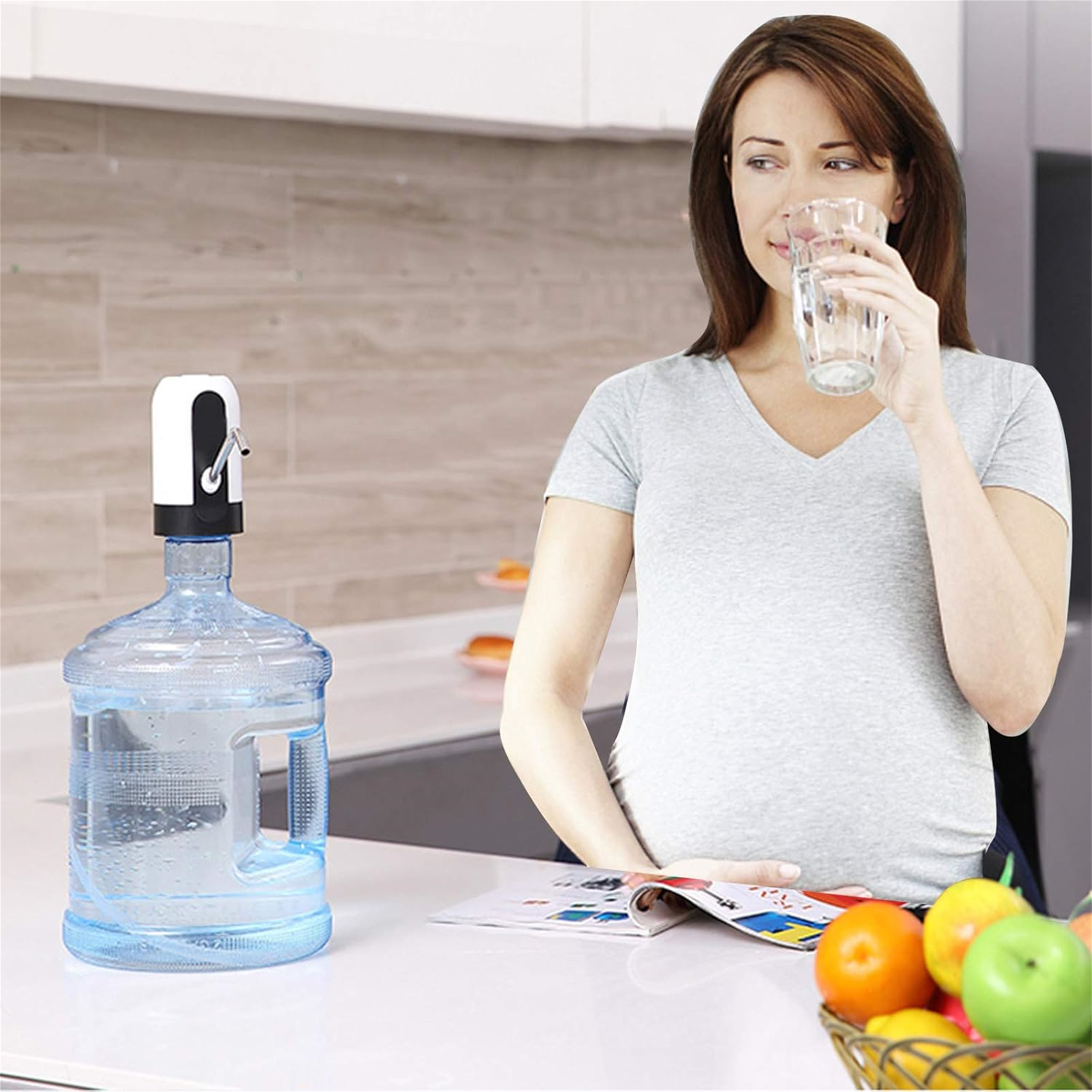 Eine Frau trinkt Wasser aus einem Glas. Links ein Wasserspender, rechts ein Obstkorb.