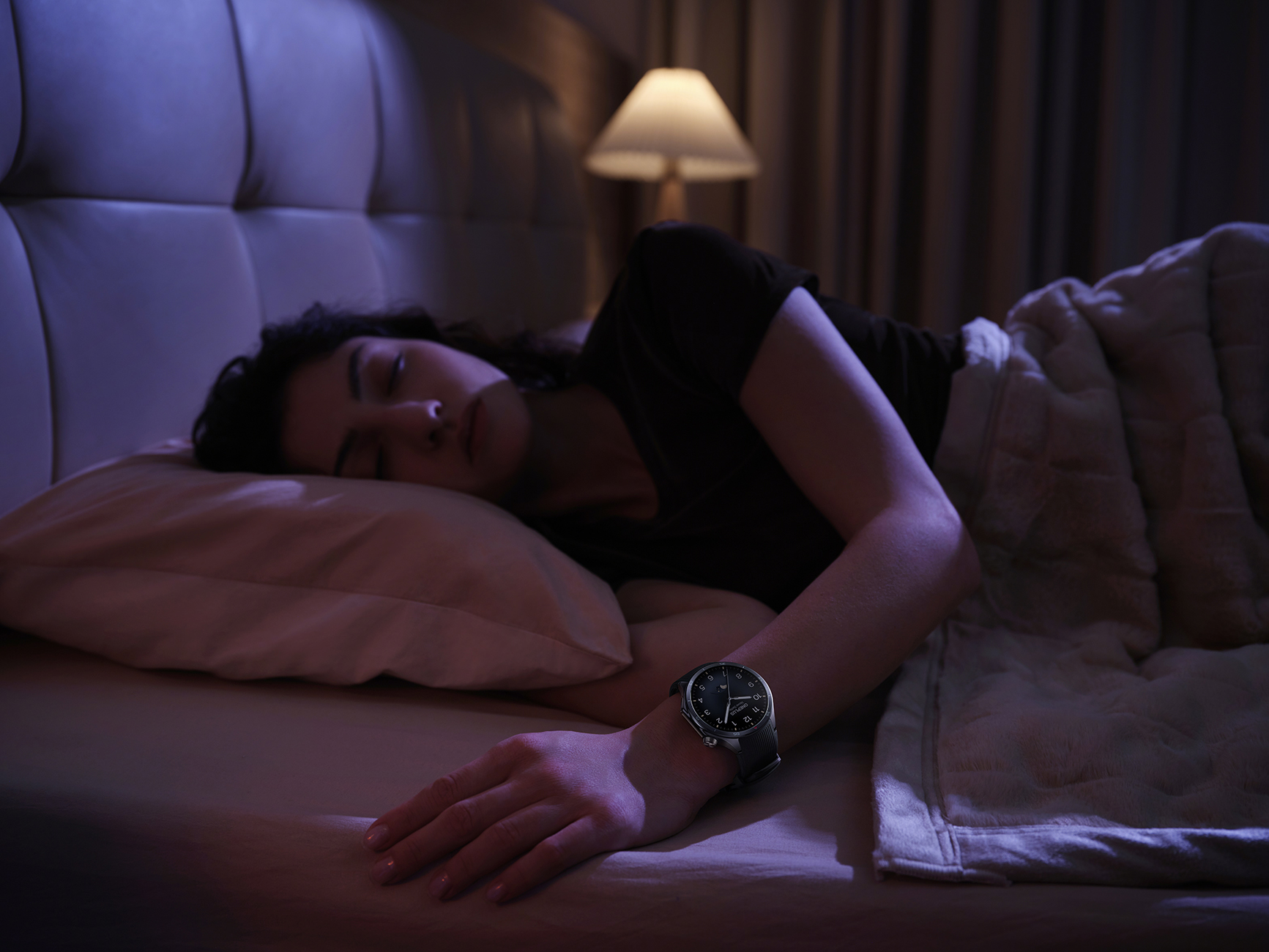 Kobieta śpi w łóżku, ma na sobie zegarek. W tle zapalona lampa. Scena jest słabo oświetlona.