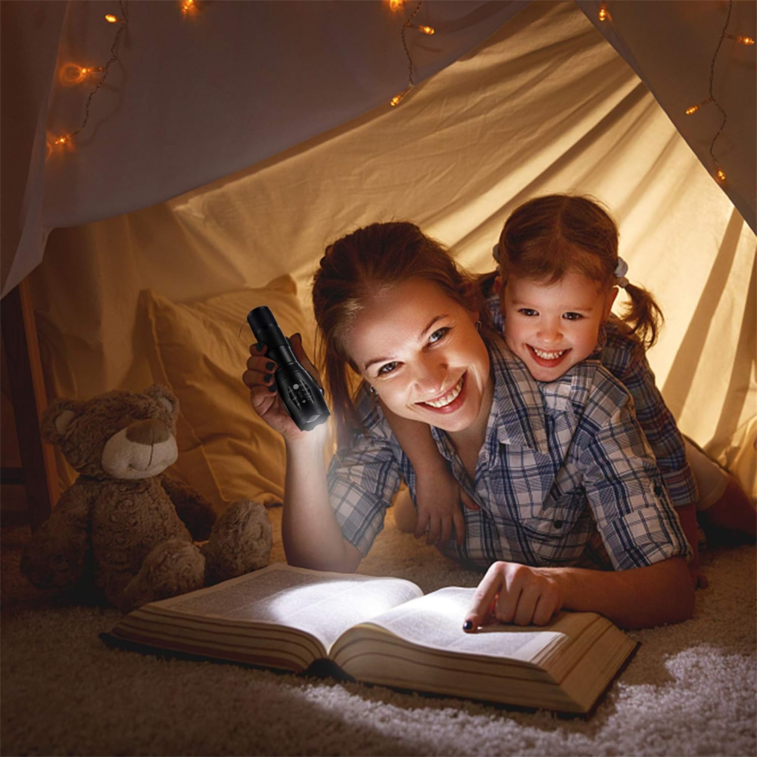Eine Frau und ein Kind lesen in einem Zelt mit einer Taschenlampe ein Buch. Ein Teddybär ist ebenfalls vorhanden.