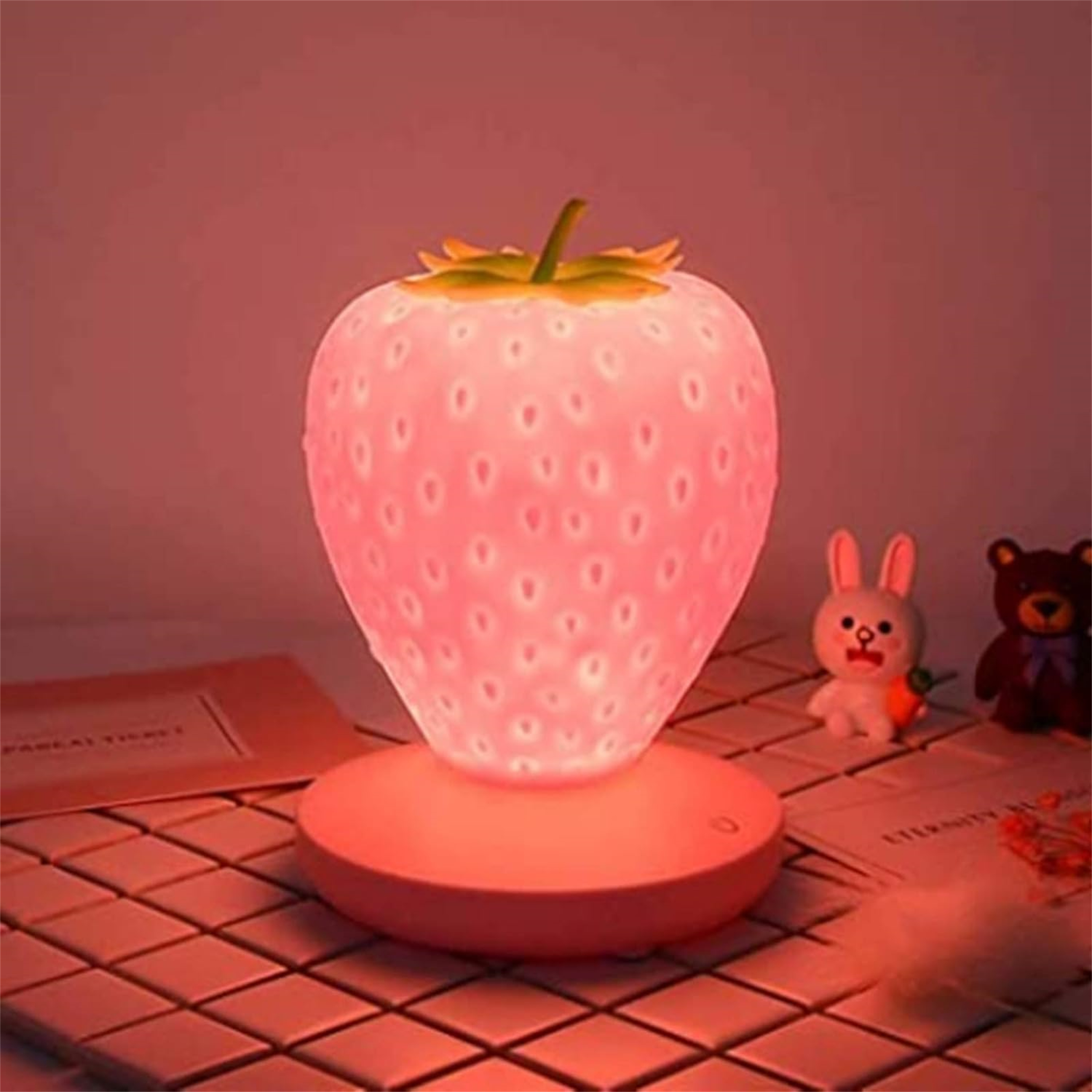 Ein rosa erdbeerförmiges Nachtlicht steht auf einem Tisch. Ein Kaninchen und ein Bär-Spielzeug sind in der Nähe. Der Hintergrund ist rosa.
