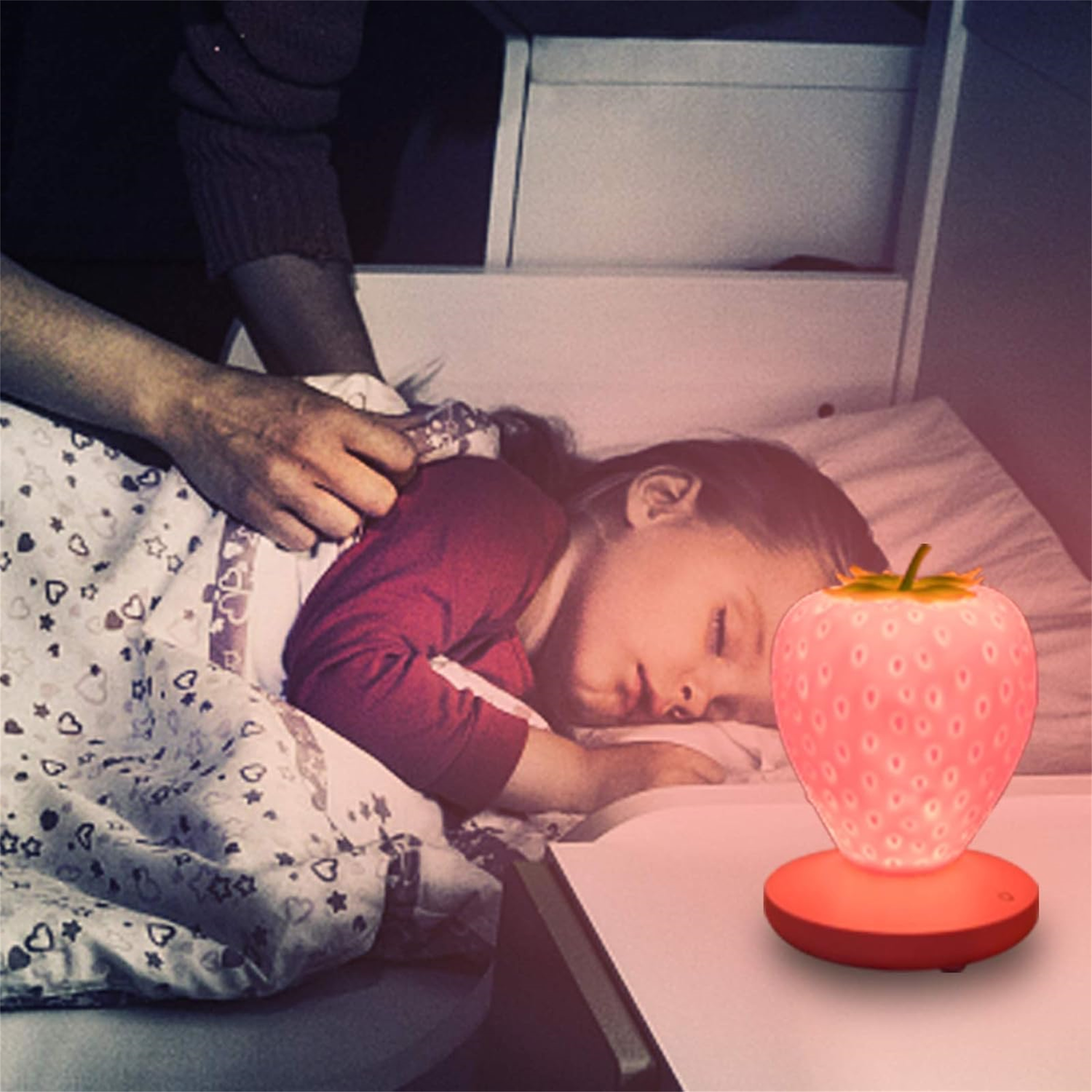 Ein schlafendes Kind liegt im Bett. Eine erdbeerförmige Lampe beleuchtet die Szene. Eine Hand richtet die Decke.