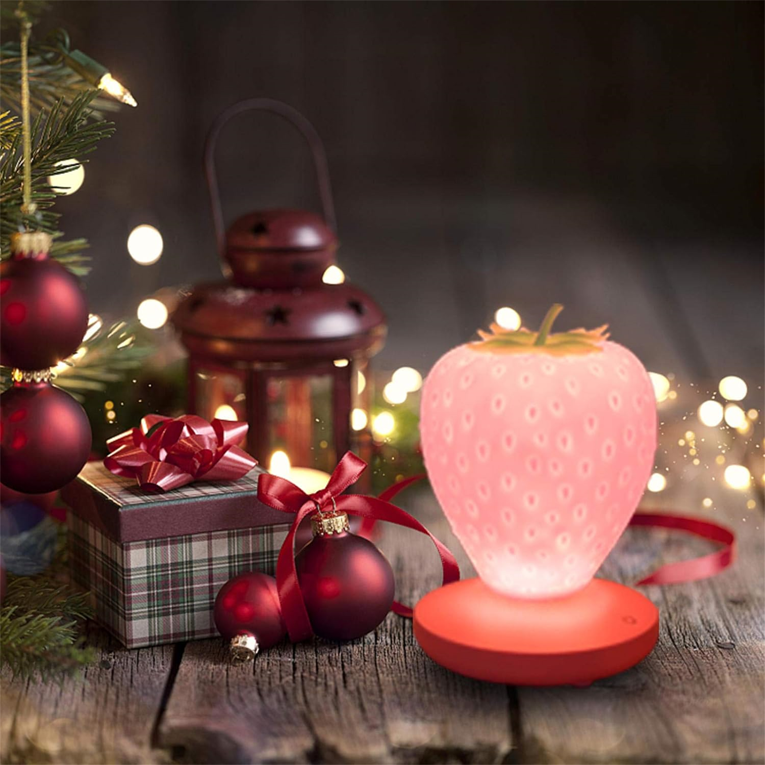 Eine erdbeerförmige Lampe leuchtet auf einem Holztisch mit Weihnachtsdekorationen. Eine Laterne und eine Geschenkbox sind im Hintergrund.