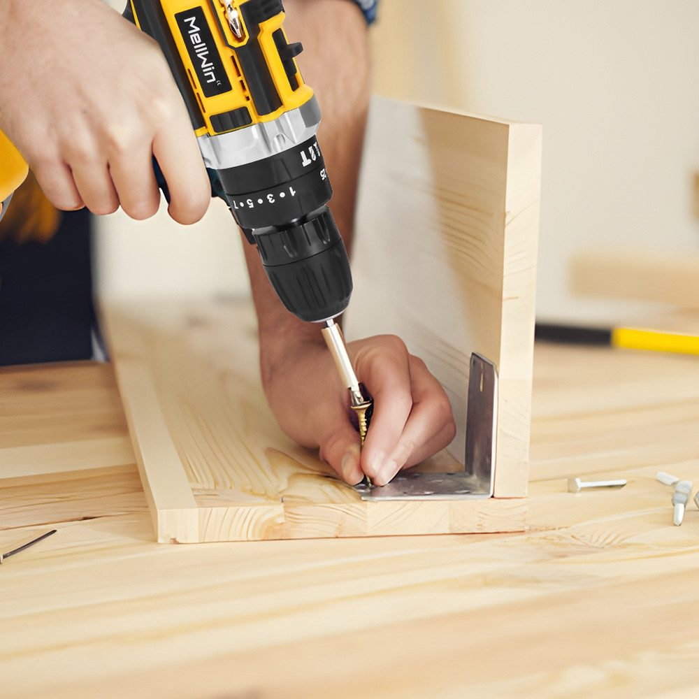 Eine Person benutzt einen gelben Bohrer, um eine Schraube in ein Holzbrett zu schrauben. Eine Metallhalterung befindet sich in der Ecke. Schrauben und Werkzeuge liegen auf dem Tisch.