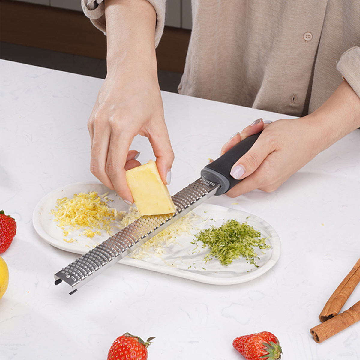 Hände reiben einen gelben Käseblock. Geriebene Schale und Käse auf einem weißen Teller. Erdbeeren, Zitrone und Zimtstangen liegen drumherum.
