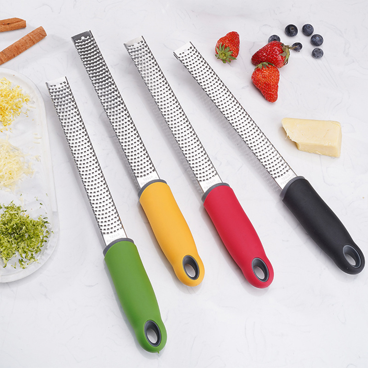 Vier Reiben mit bunten Griffen, Erdbeeren, Blaubeeren und Käse auf weißer Oberfläche.