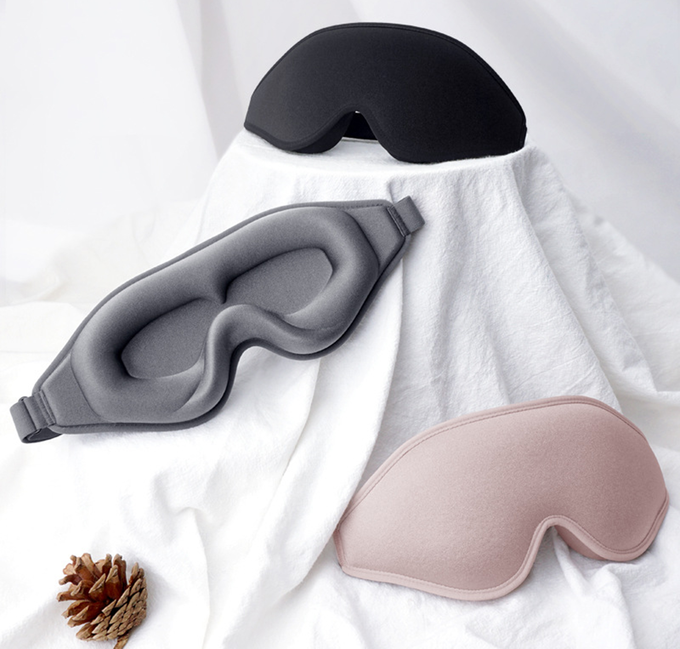 Drei Augenmasken in Schwarz, Grau und Rosa liegen auf weißem Stoff. Unten links eine Kiefernzapfen.