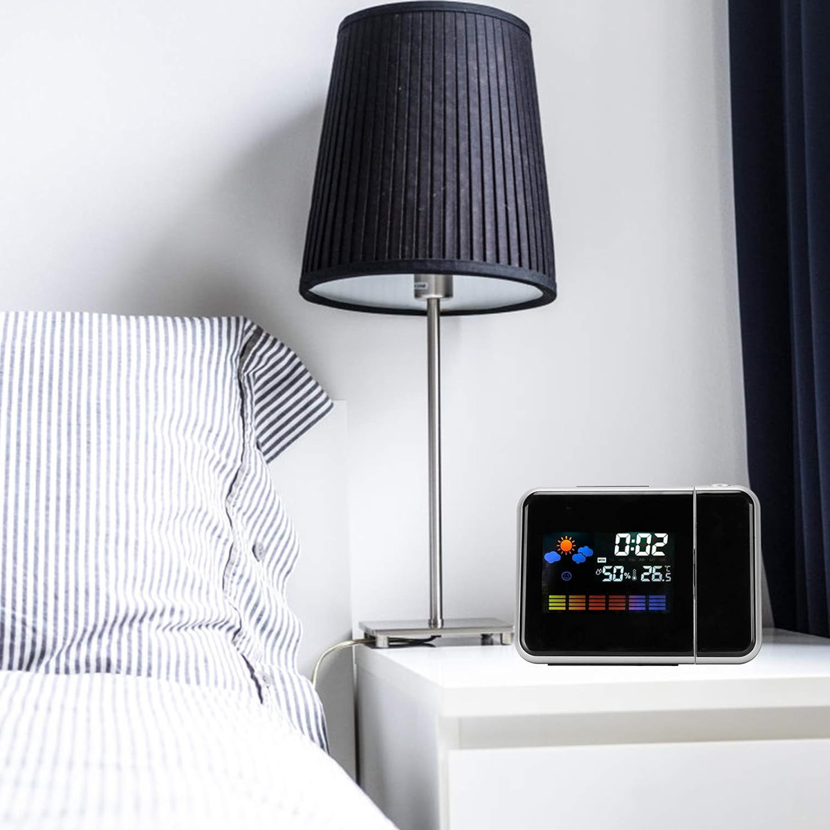 Ein Nachttisch mit einer Digitaluhr, die Zeit und Wetter anzeigt. Eine schwarze Lampe steht auf dem Tisch, ein gestreiftes Kissen und Vorhänge sind sichtbar.