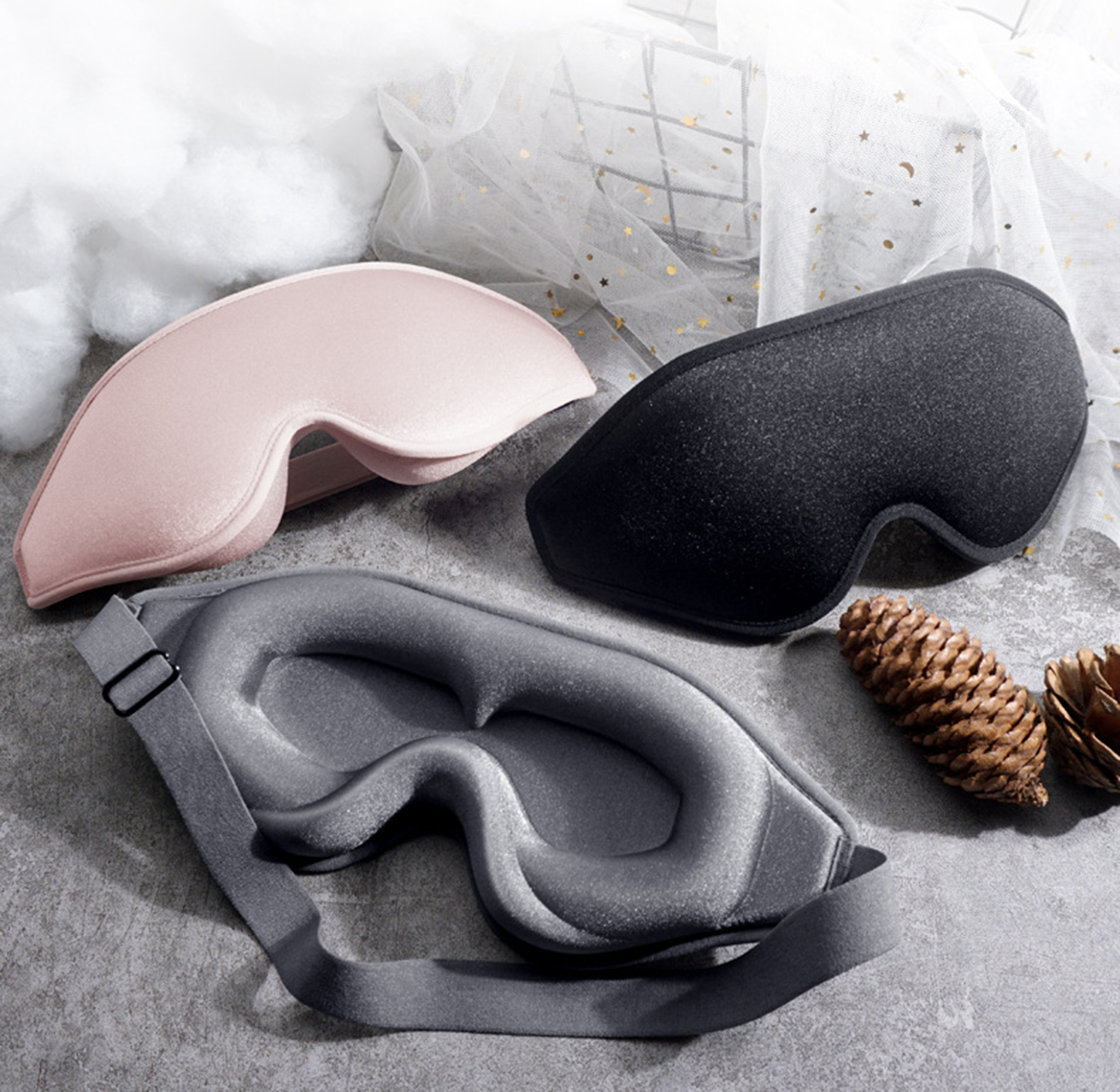 Drei Schlafmasken: rosa, schwarz und grau. Eine Kiefernzapfen und ein weißes Tuch mit goldenen Sternen im Hintergrund.