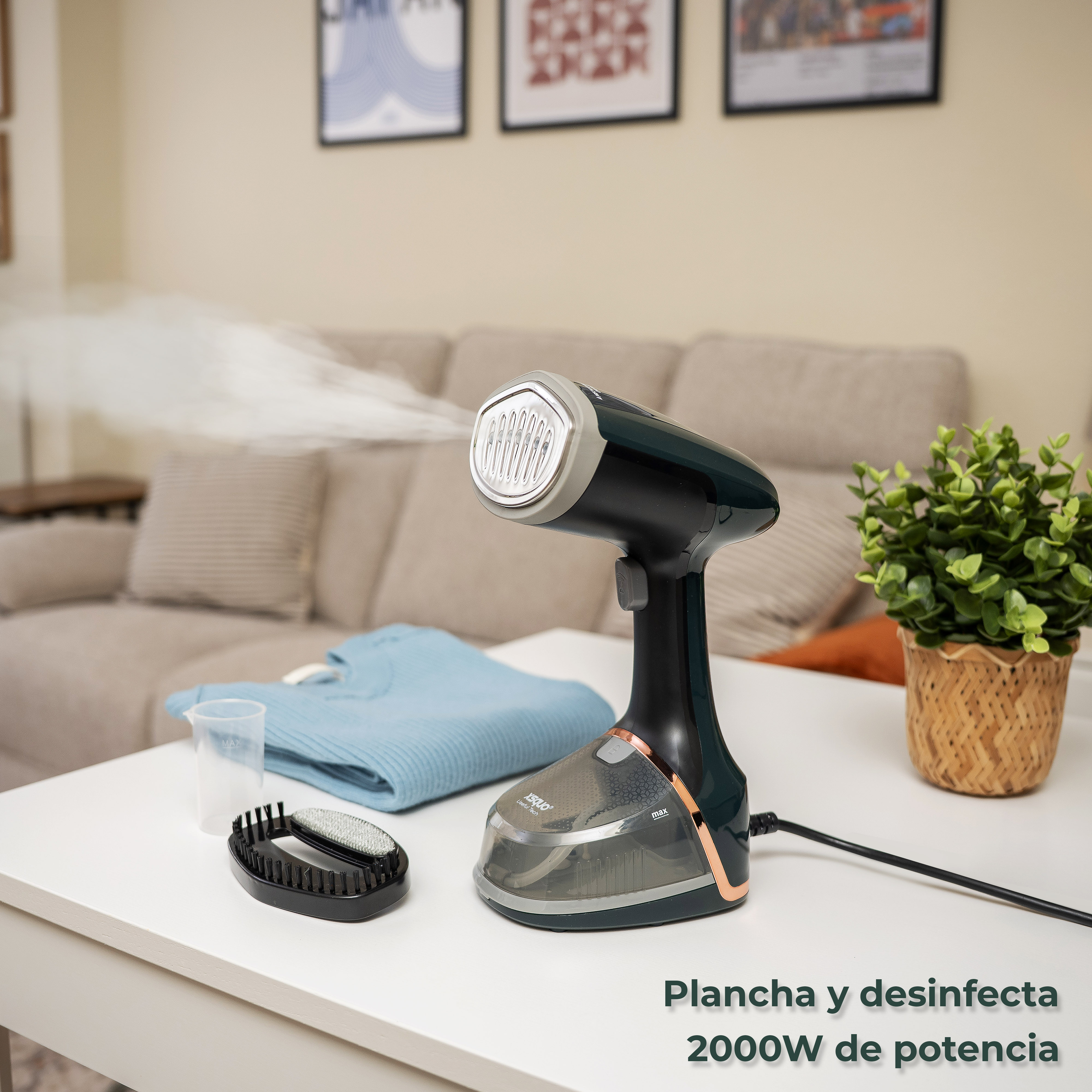 Un vaporizador de mano negro y verde azulado con vapor, ropa y accesorios sobre una mesa blanca. Hay una planta a la derecha.