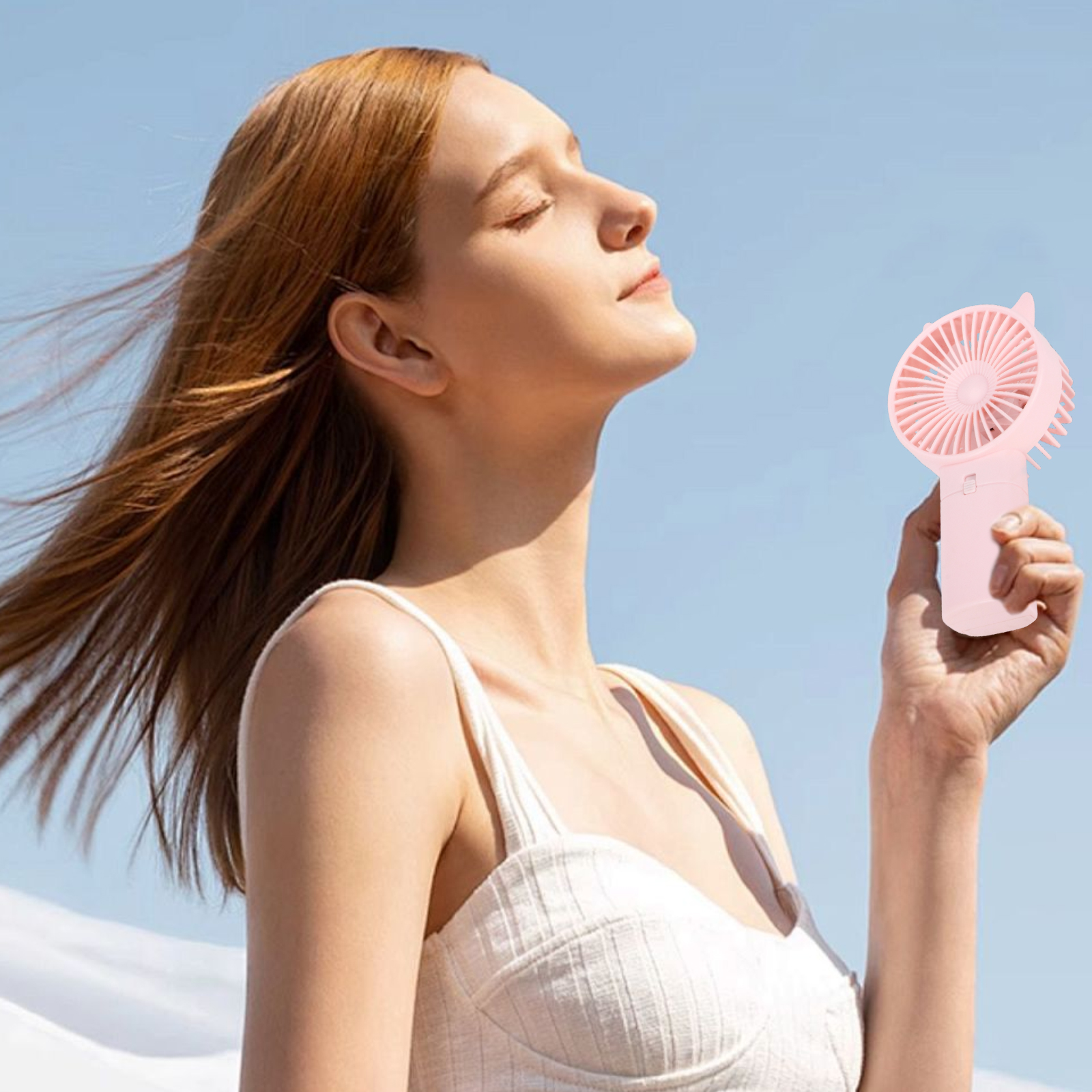 Eine Frau hält einen rosa Fächer, die Augen geschlossen, vor blauem Himmel. Ihr Haar weht im Wind. Sie trägt ein weißes Oberteil.