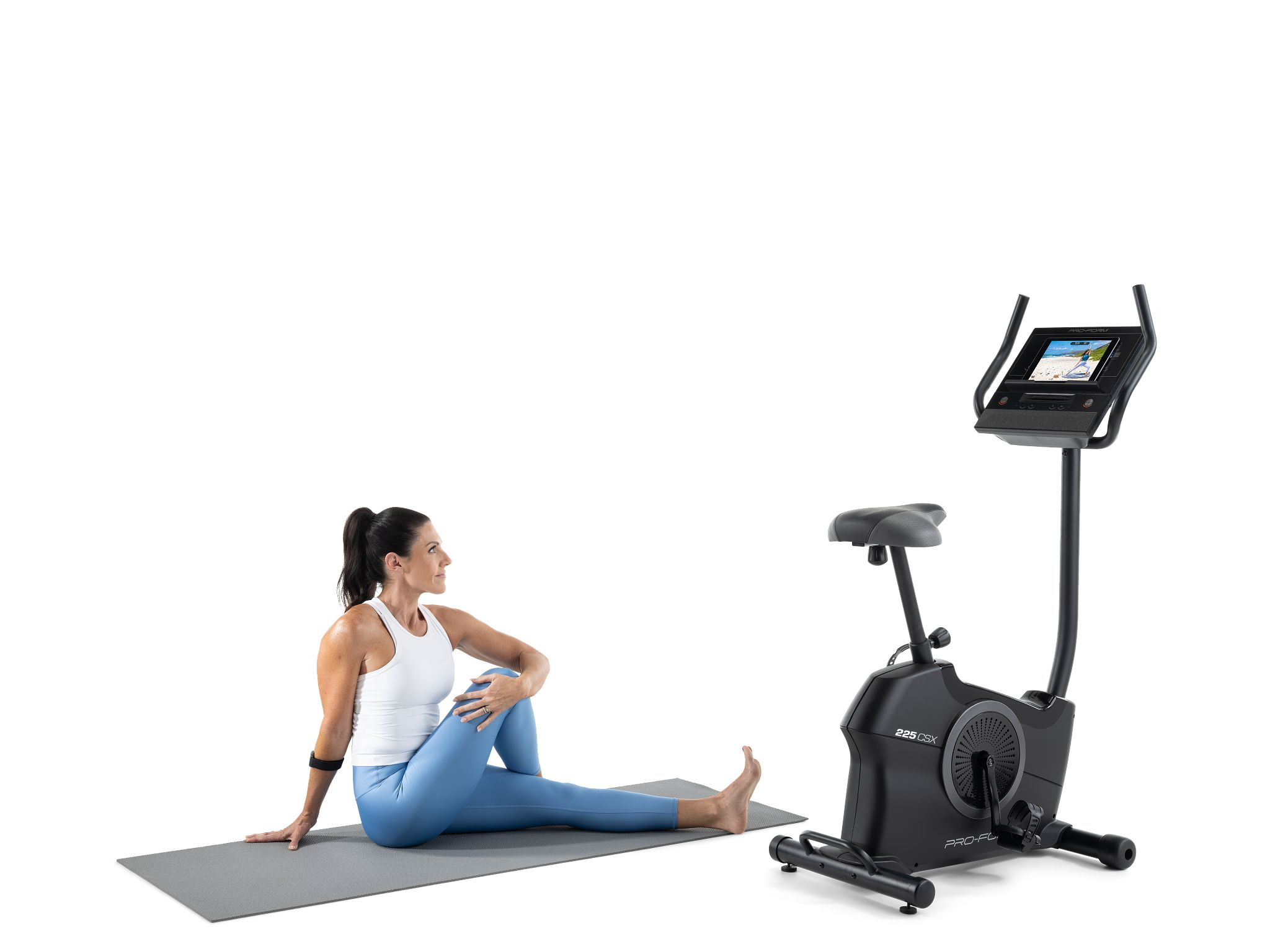 Eine Frau dehnt sich auf einer Matte neben einem Heimtrainer. Sie trägt ein weißes Oberteil und blaue Leggings. Das Fahrrad ist schwarz.