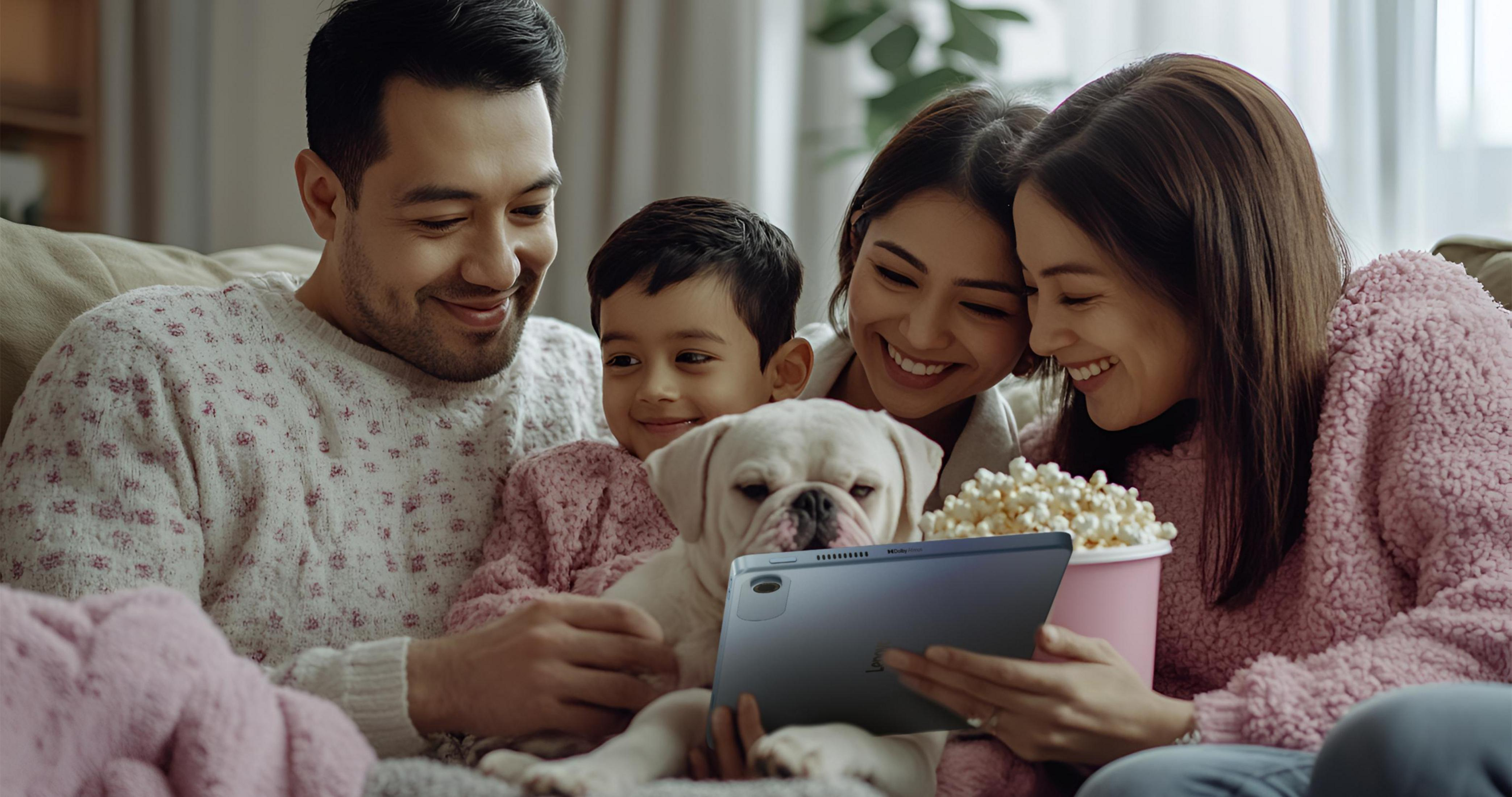 Una famiglia guarda un tablet con un cane. Stanno sorridendo. Un secchio di popcorn è vicino.