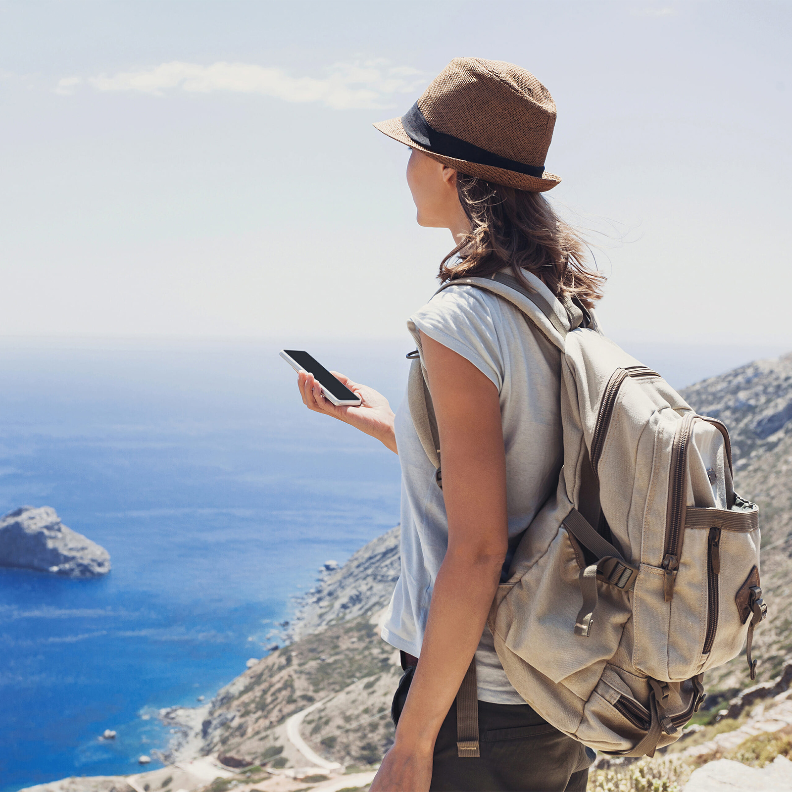 Frau mit Hut und Rucksack, hält ein Telefon, mit Blick auf ein Meer. Blaues Wasser, Himmel und Berge sichtbar.