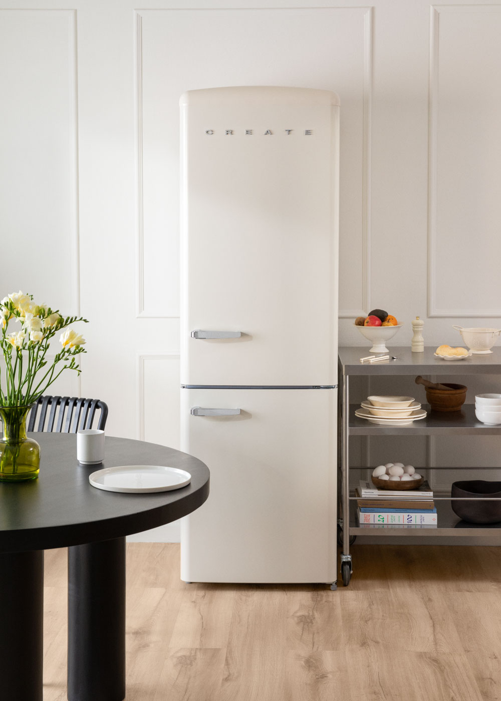 Una escena de cocina. Una nevera blanca se encuentra junto a una estantería de acero inoxidable. Una mesa negra y un jarrón con flores están en primer plano.