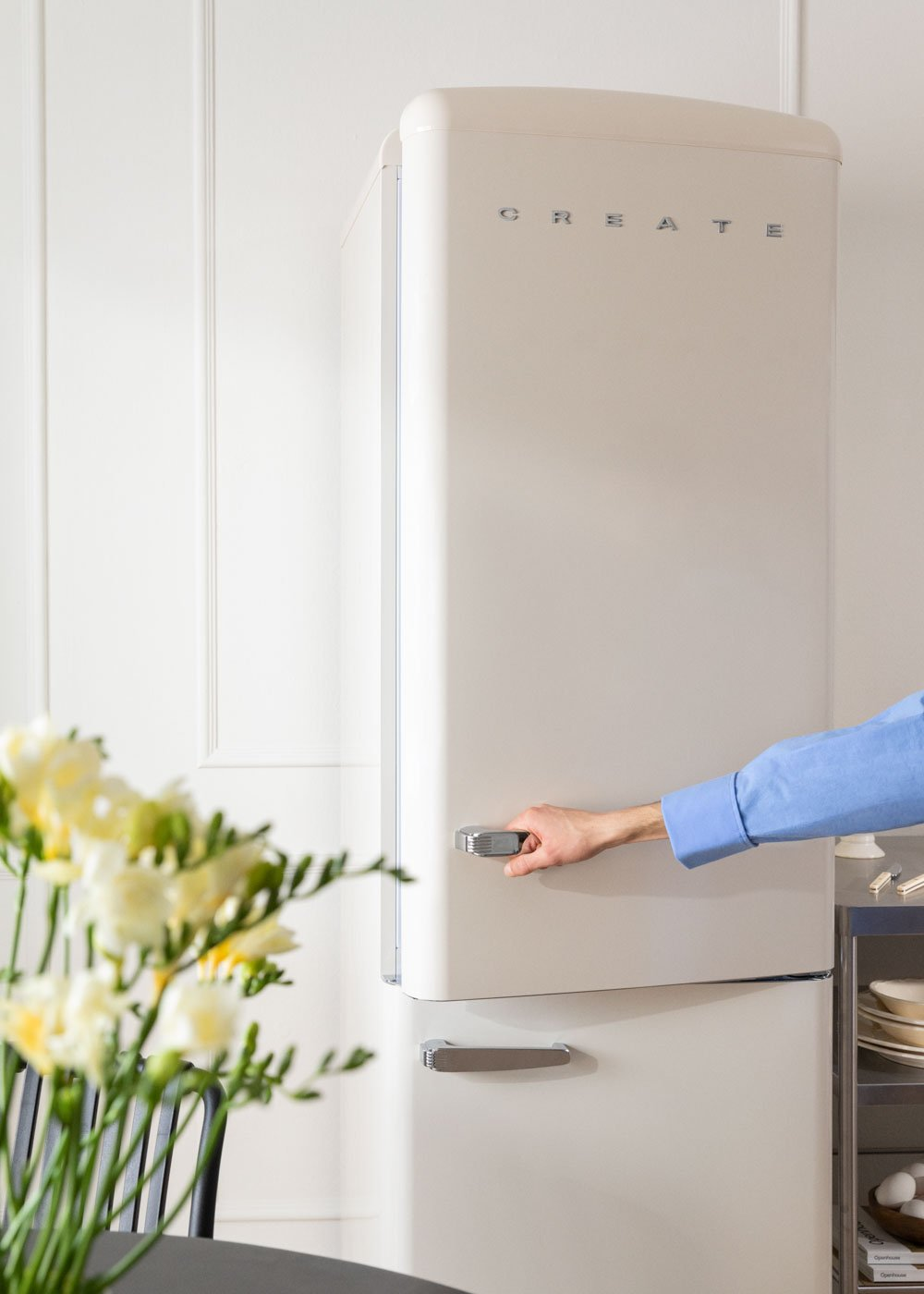Una persona abre un refrigerador color crema. La palabra 'CREATE' está en el refrigerador. Un ramo de flores está a la izquierda.