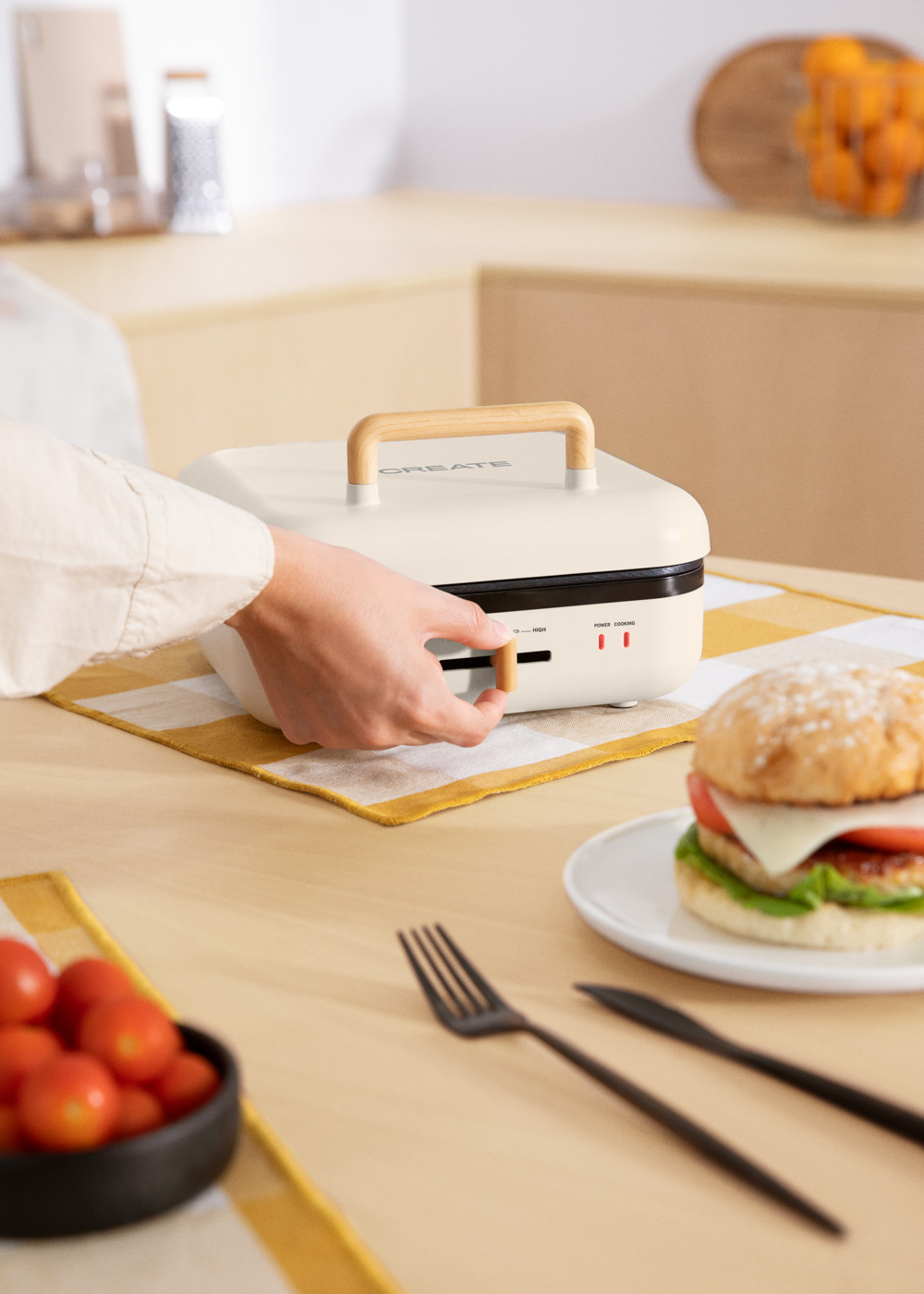 Una persona usando una sandwichera en una mesa. Una hamburguesa y tomates están en la mesa. También hay un tenedor y un cuchillo.