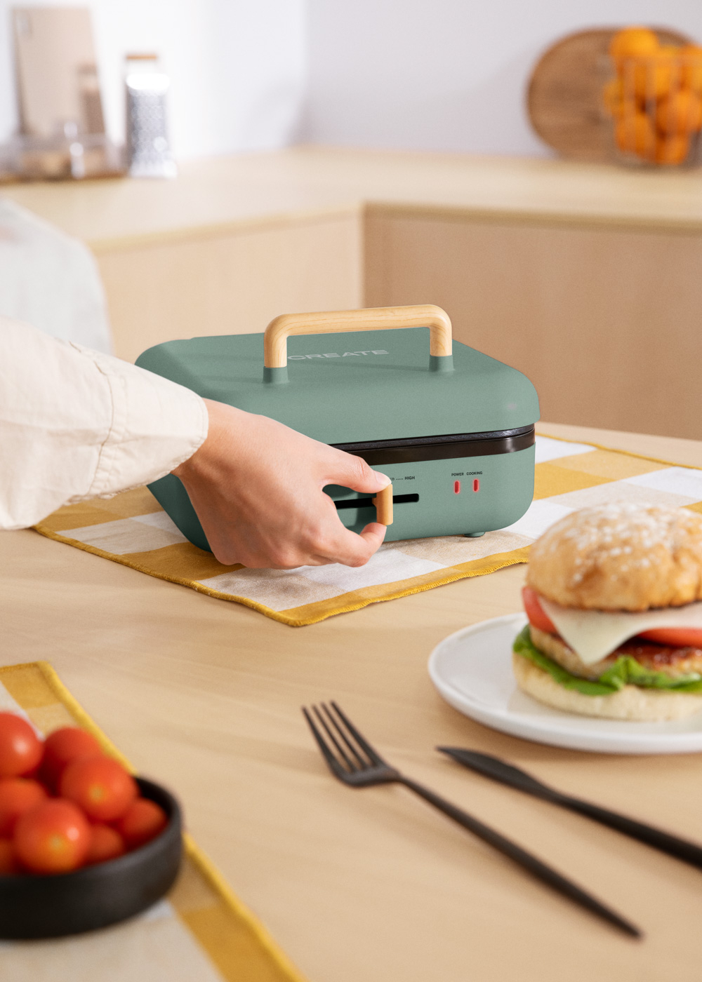 Una persona ajusta una sandwichera verde. Una hamburguesa está en un plato. Tomates, un tenedor y un cuchillo están sobre la mesa.