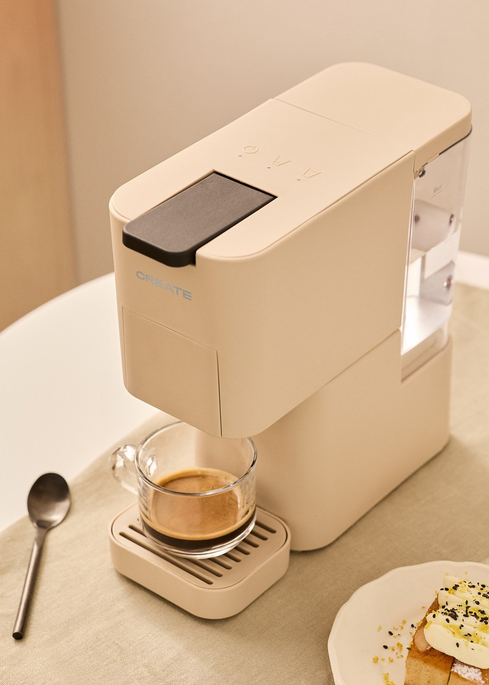 Una cafetera beige con un vaso de espresso. Una cuchara y pastel en la mesa. La máquina dice 'CREATE'.