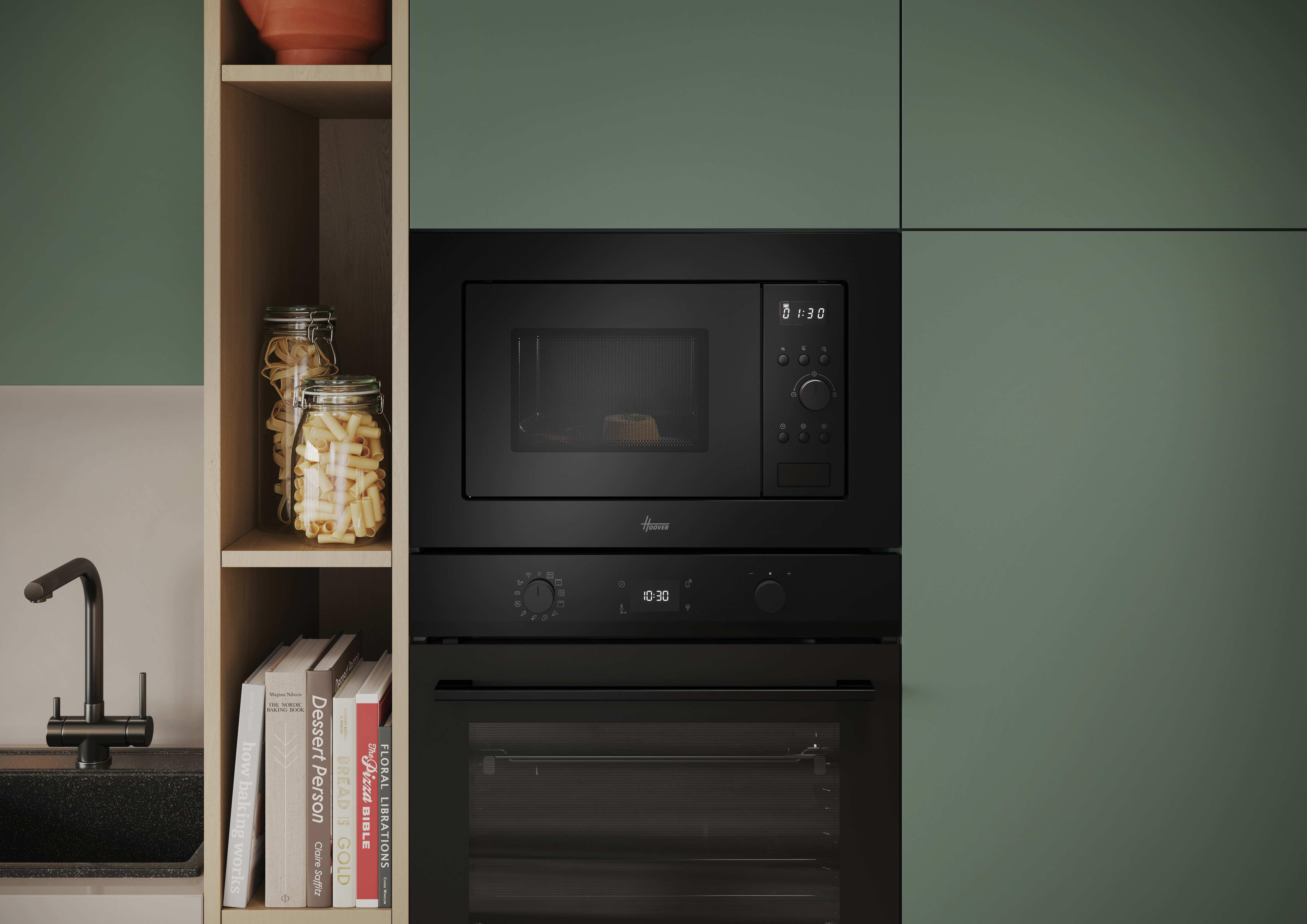 Una escena de cocina con horno y microondas negros. Estantes con pasta y libros de cocina a la izquierda. Armarios verdes.