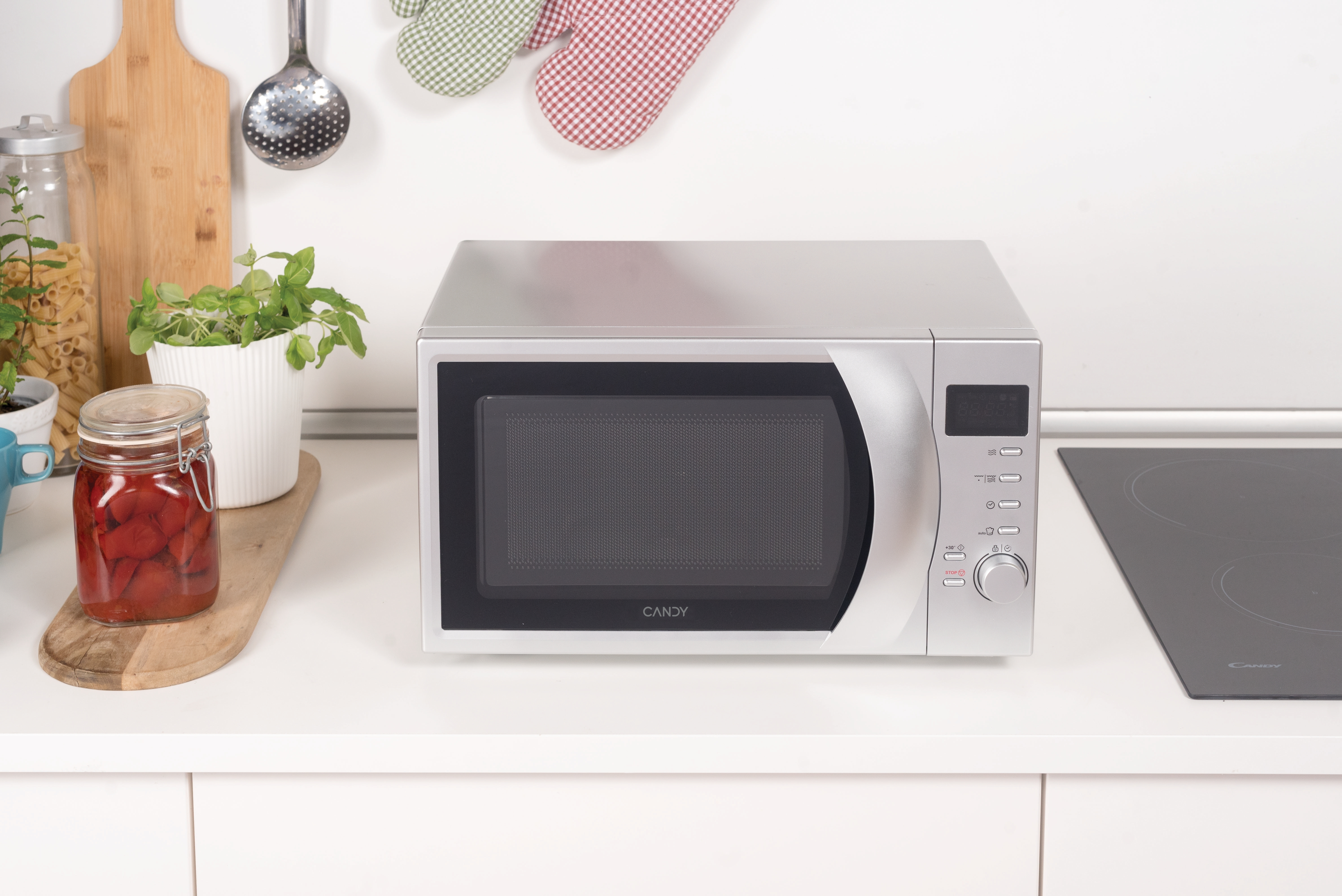 Un forno a microonde argentato su un piano di lavoro bianco. Dietro, utensili da cucina e piante.