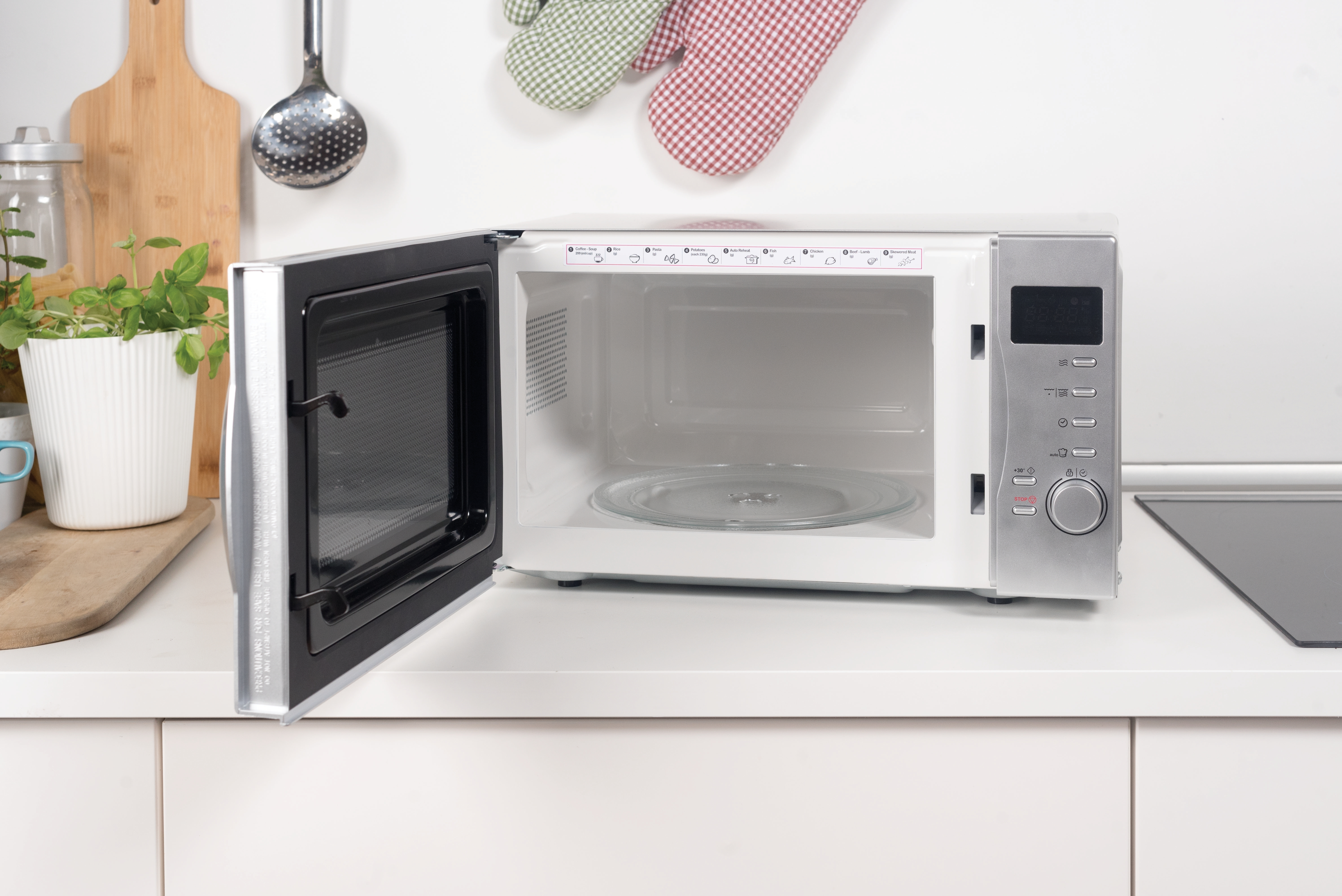 Un forno a microonde aperto su un bancone bianco. Nel retro sono visibili un tagliere, erbe e utensili.