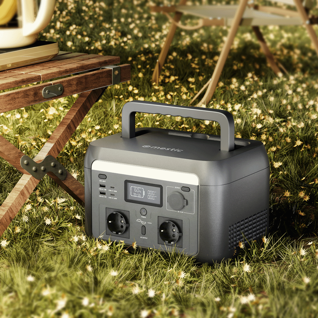 A portable power station sits on grass, near a camping table. The station is gray with a handle. Camping chairs are visible.