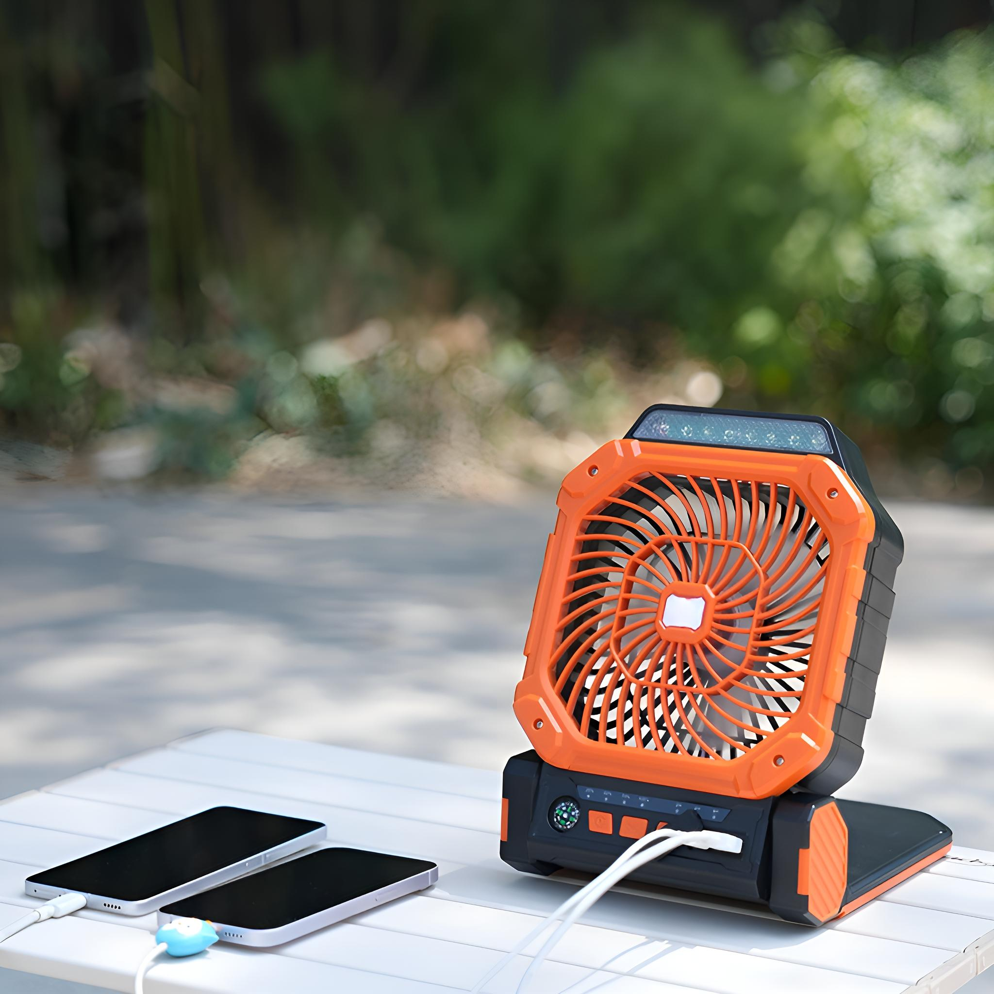 Ein orange-schwarzer Ventilator steht auf einem weißen Tisch mit zwei ladenden Telefonen. Verschwommene grüne Bäume im Hintergrund.