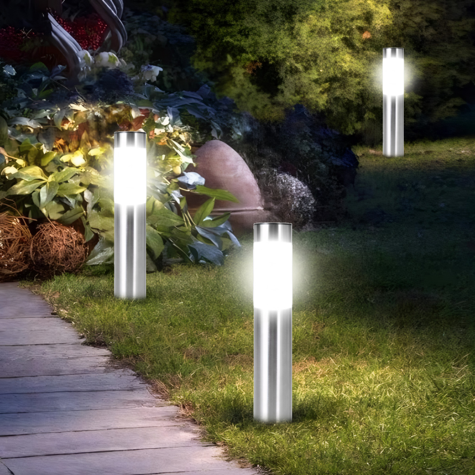 Gartenszene mit drei beleuchteten silbernen Lampen auf Gras, einem Holzweg und Laub im Hintergrund. Die Szene ist nachts beleuchtet.