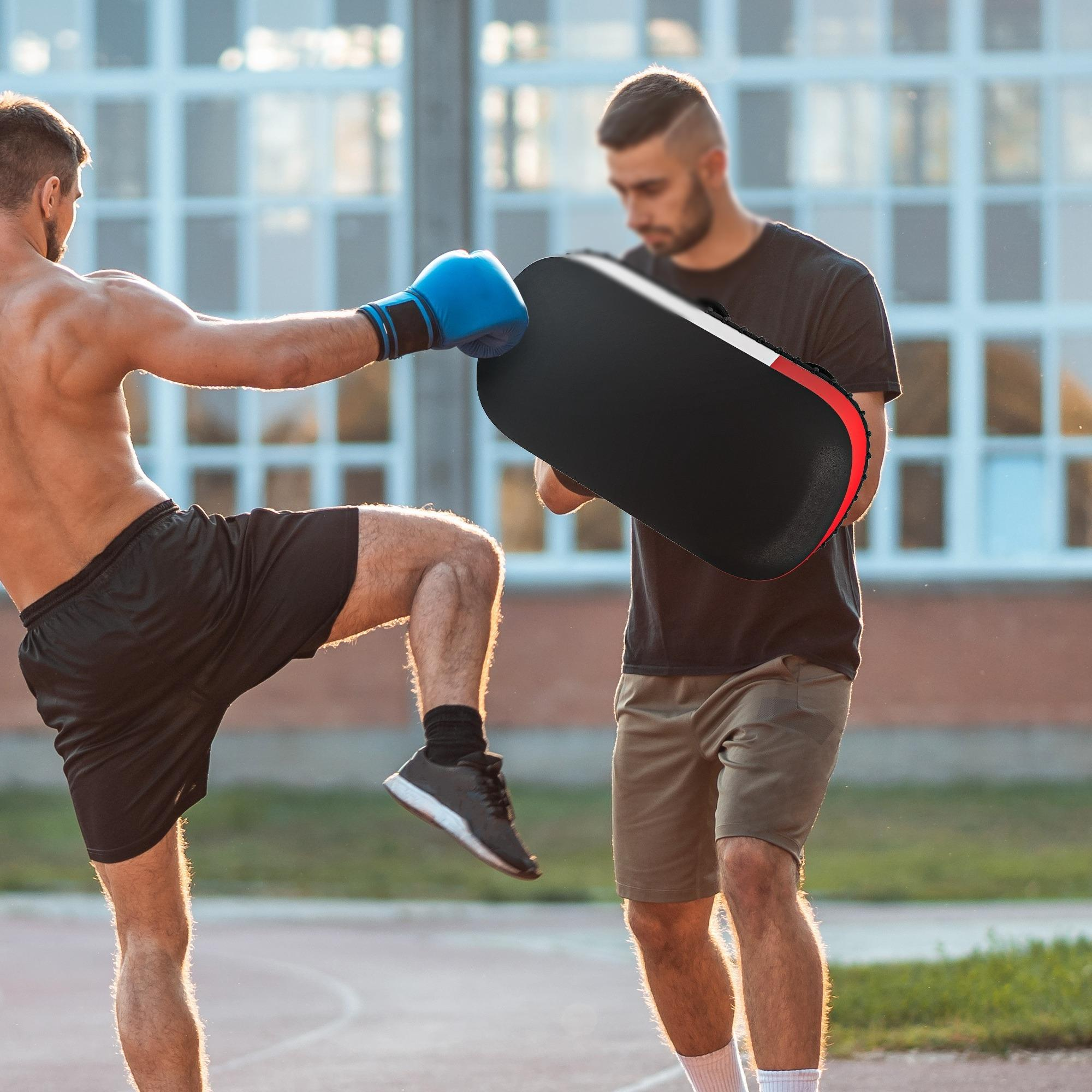 Ein Mann mit blauen Handschuhen tritt auf ein von einem anderen gehaltenes Pad. Schwarze Shorts, schwarzes Pad mit roter Umrandung.