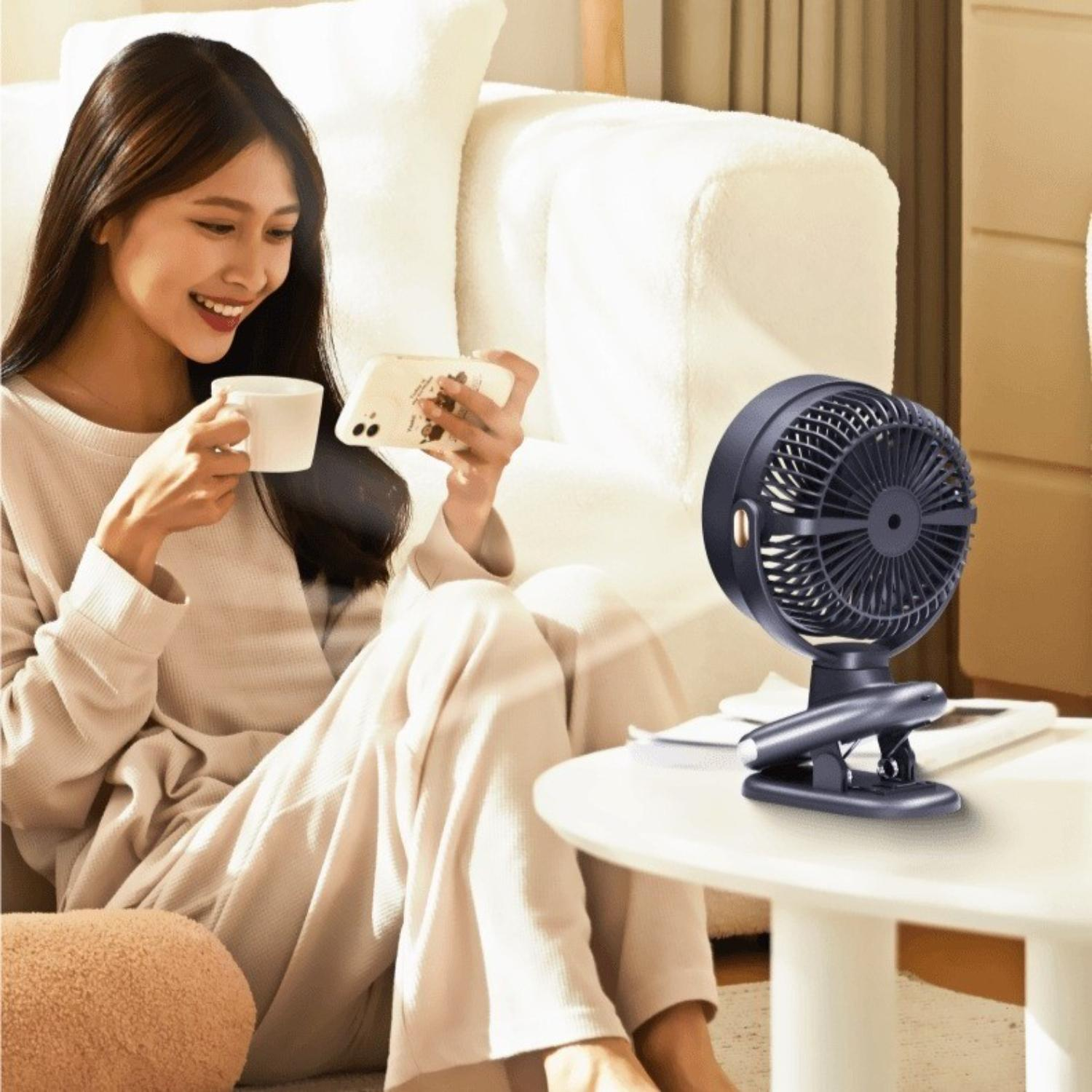 Eine Frau sitzt drinnen, benutzt ihr Telefon und hält eine Tasse. Ein kleiner Ventilator steht auf einem Tisch. Hintergrund ist ein Sofa und ein kleines Kissen.