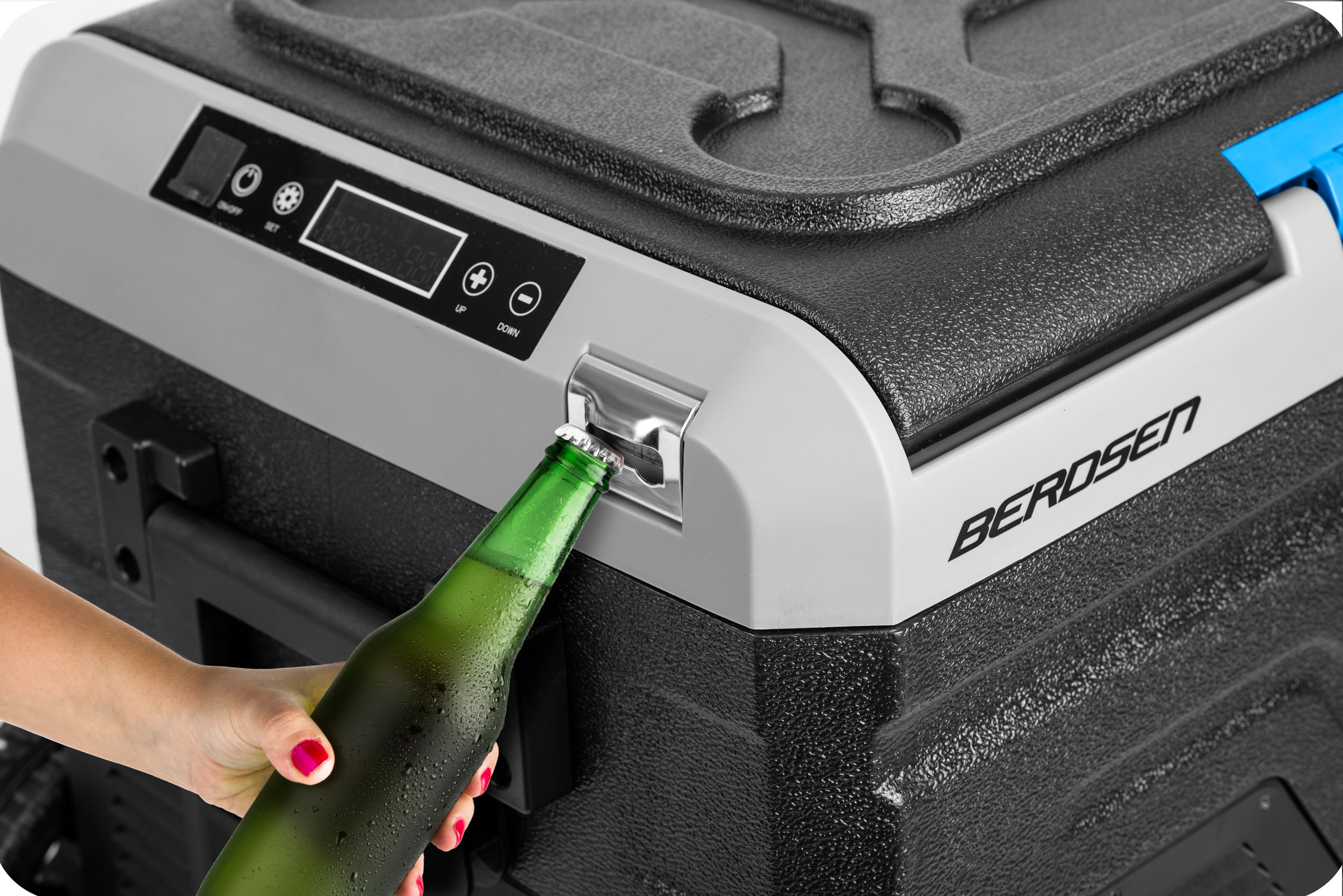 Zielona butelka piwa jest otwierana. Znajduje się obok czarno-szarej lodówki z cyfrowym wyświetlaczem. Ręka trzyma butelkę.