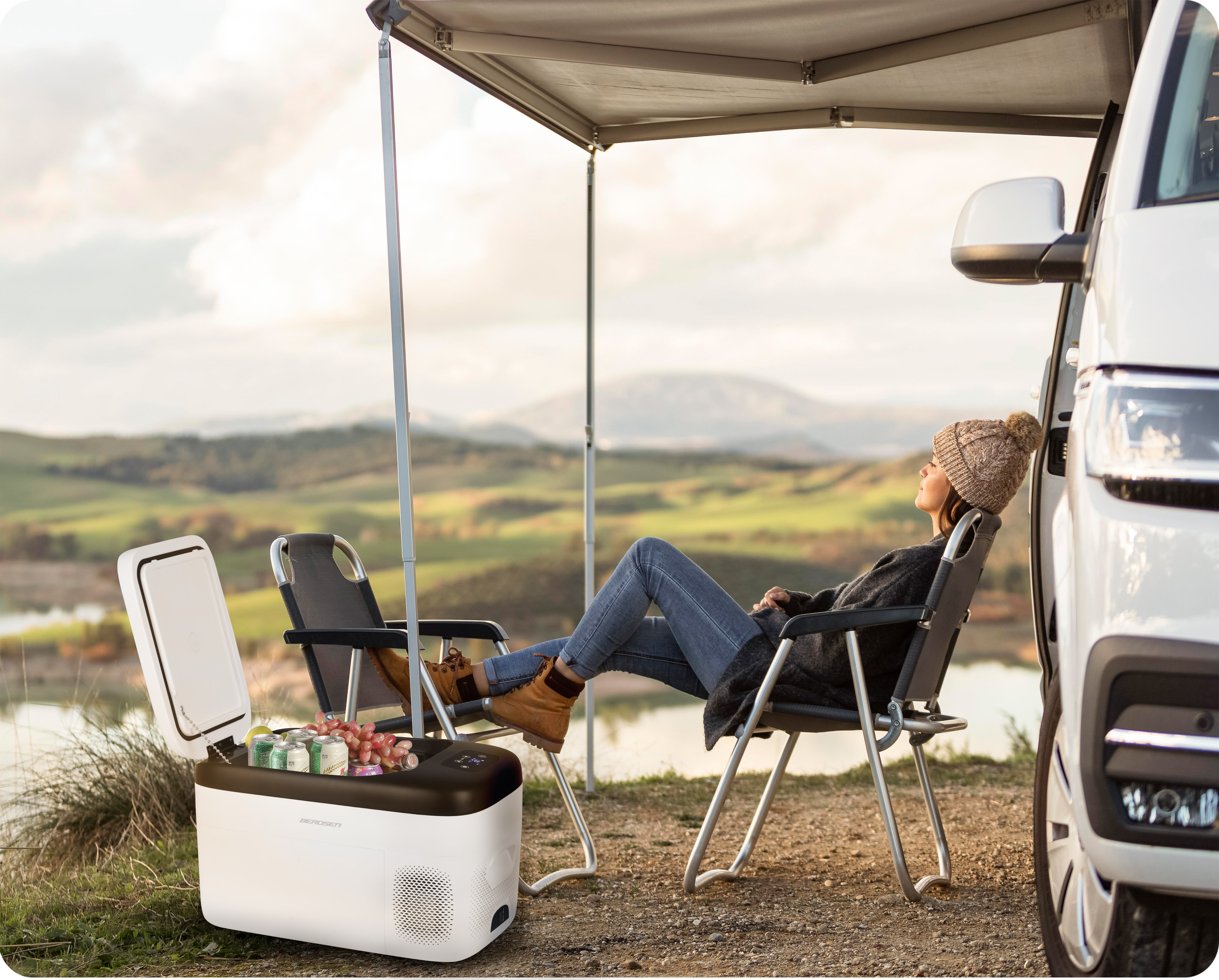 Kobieta relaksuje się na krześle kempingowym, w pobliżu vana. Otwarta lodówka z napojami. Góry w tle.