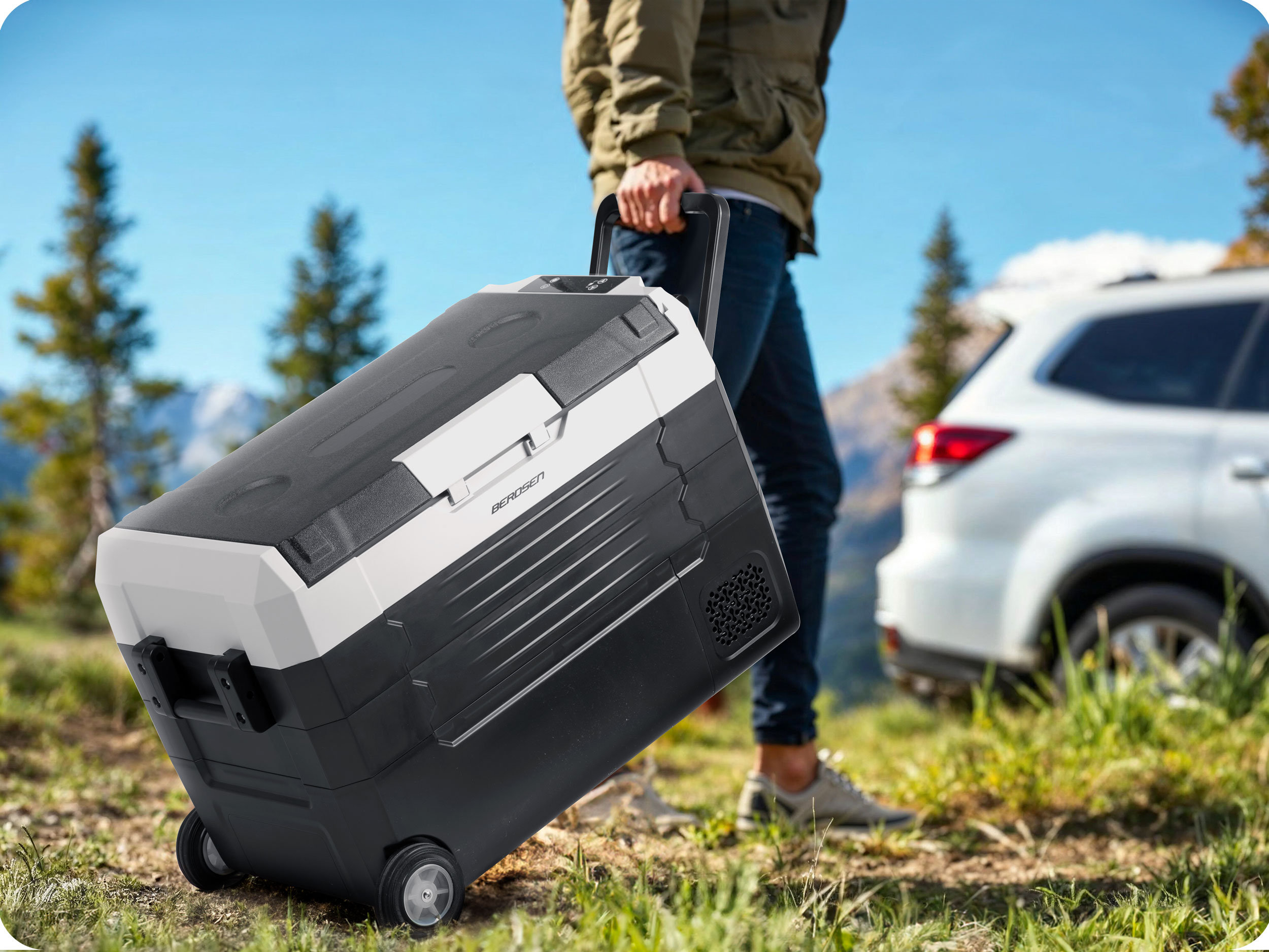 Osoba ciągnie czarno-białą chłodziarkę na kółkach na zewnątrz, z białym samochodem w tle.