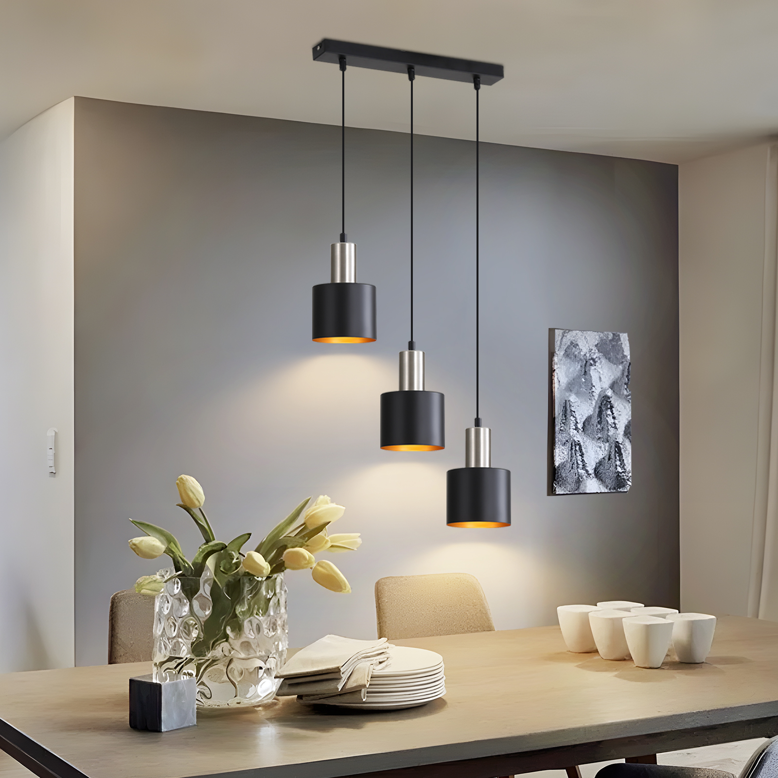 Esszimmer mit drei Pendelleuchten, Tisch mit Geschirr, Blumen und sechs weißen Tassen. Graue Wand und Kunstwerk.