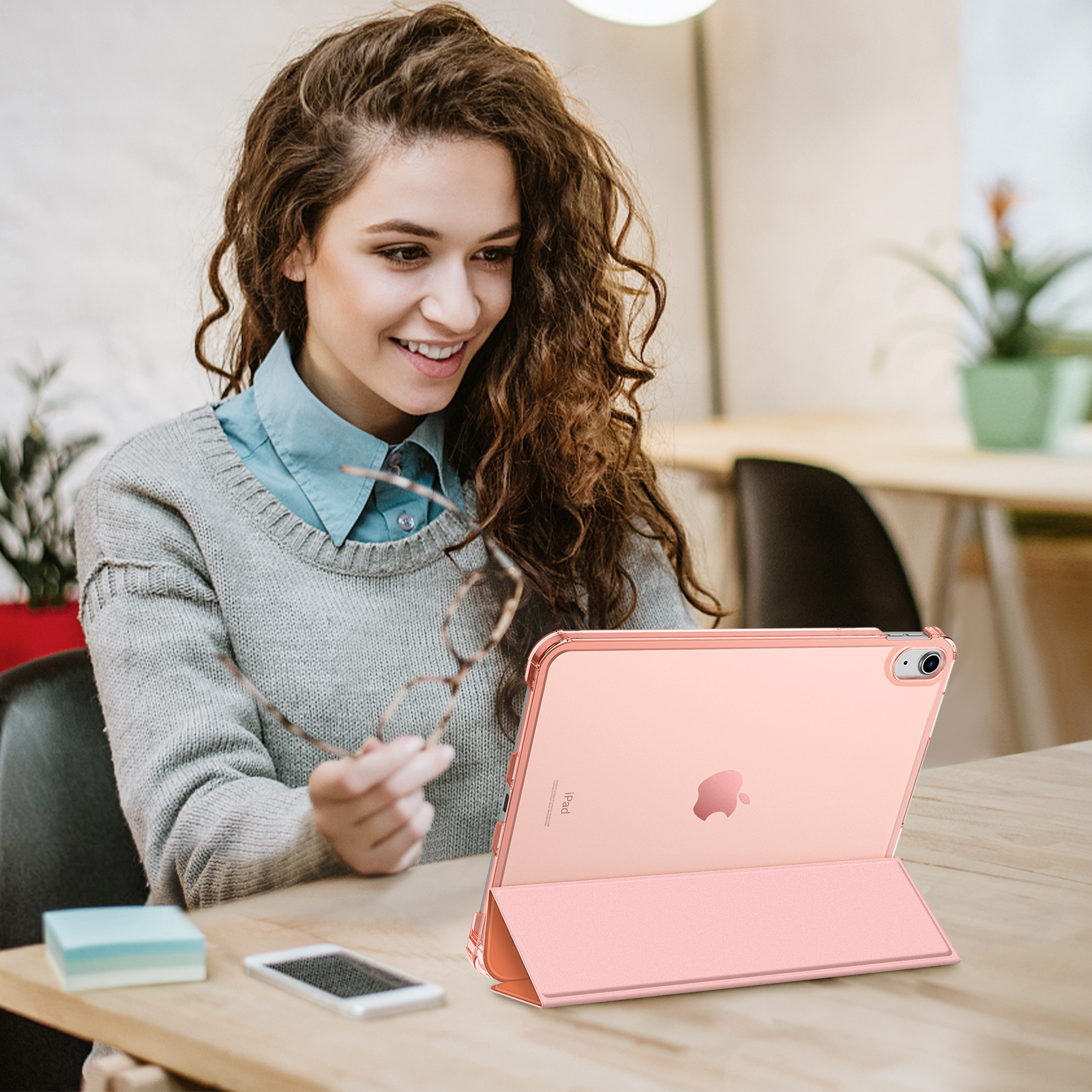 Eine lächelnde Frau hält eine Brille. Ein rosa iPad liegt auf einem Tisch. Ein Telefon und Haftnotizen sind ebenfalls auf dem Tisch.