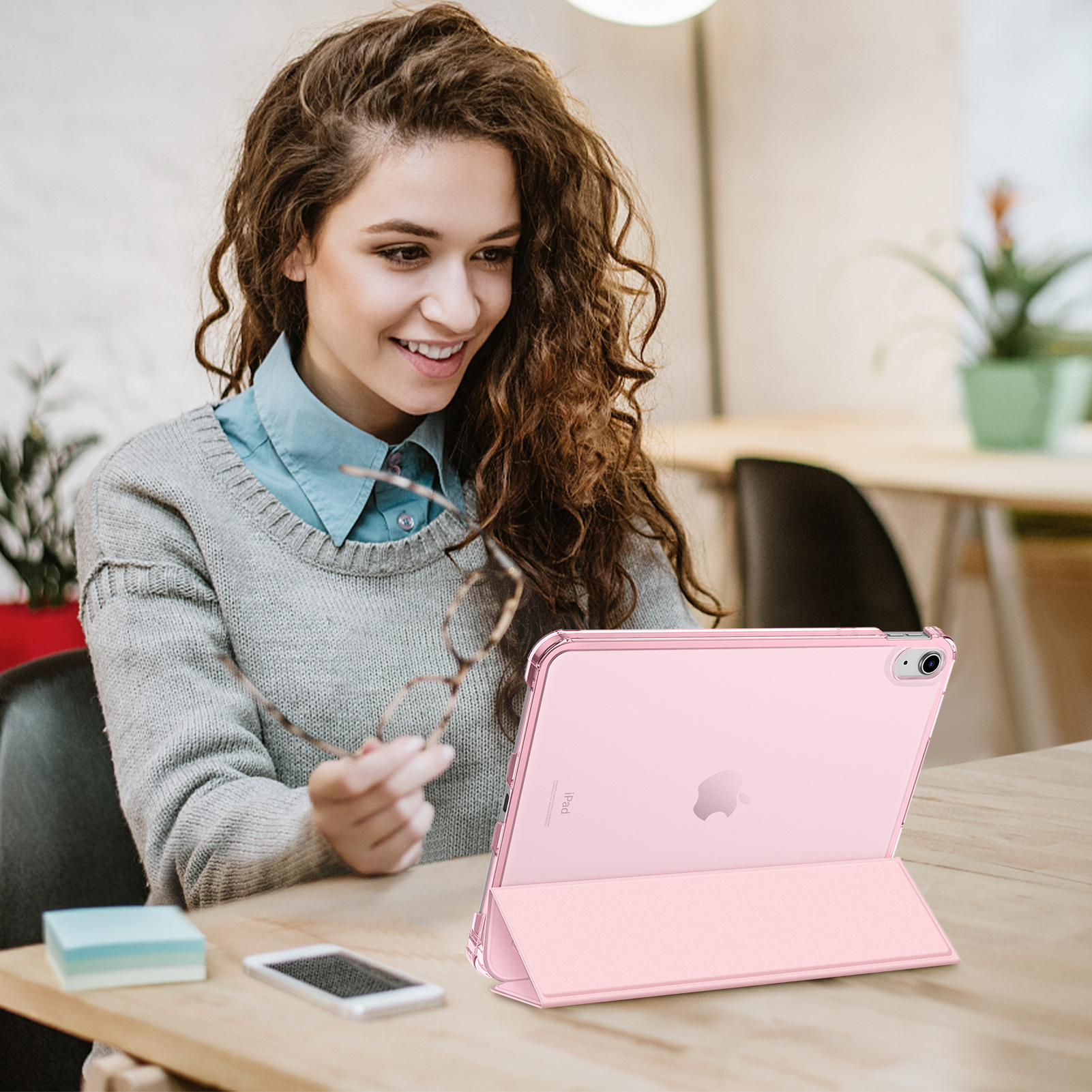 Lächelnde Frau, Brille haltend, blickt auf ein rosa iPad auf einem Holztisch. Ein Smartphone und ein Notizblock liegen ebenfalls auf dem Tisch.