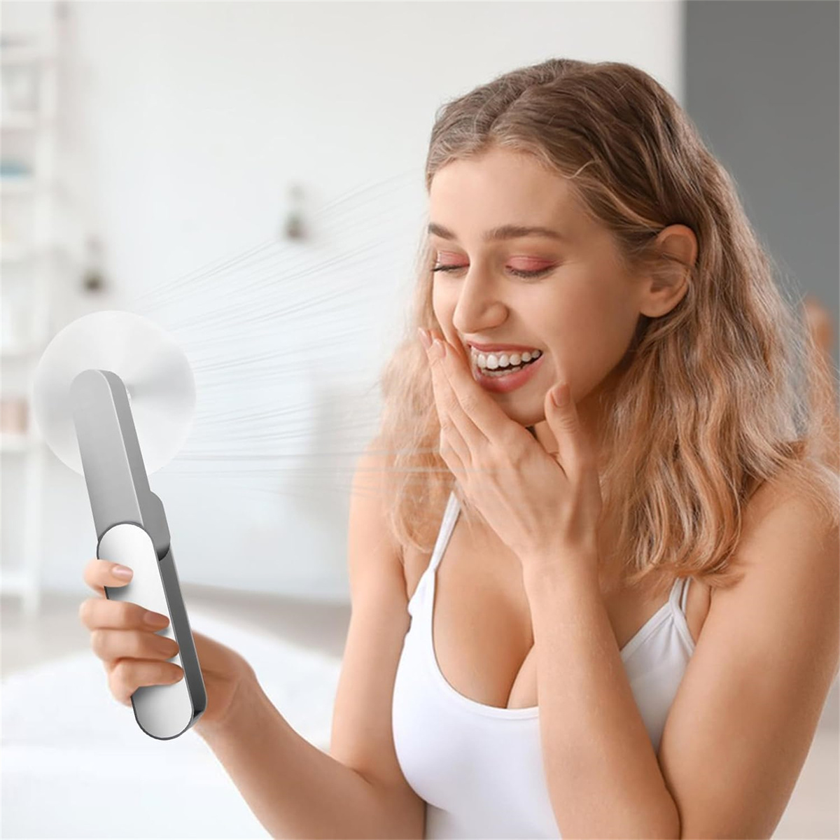 Eine Frau lächelt und hält einen kleinen Ventilator. Der Ventilator bläst ihr Luft ins Gesicht. Sie trägt ein weißes Top.