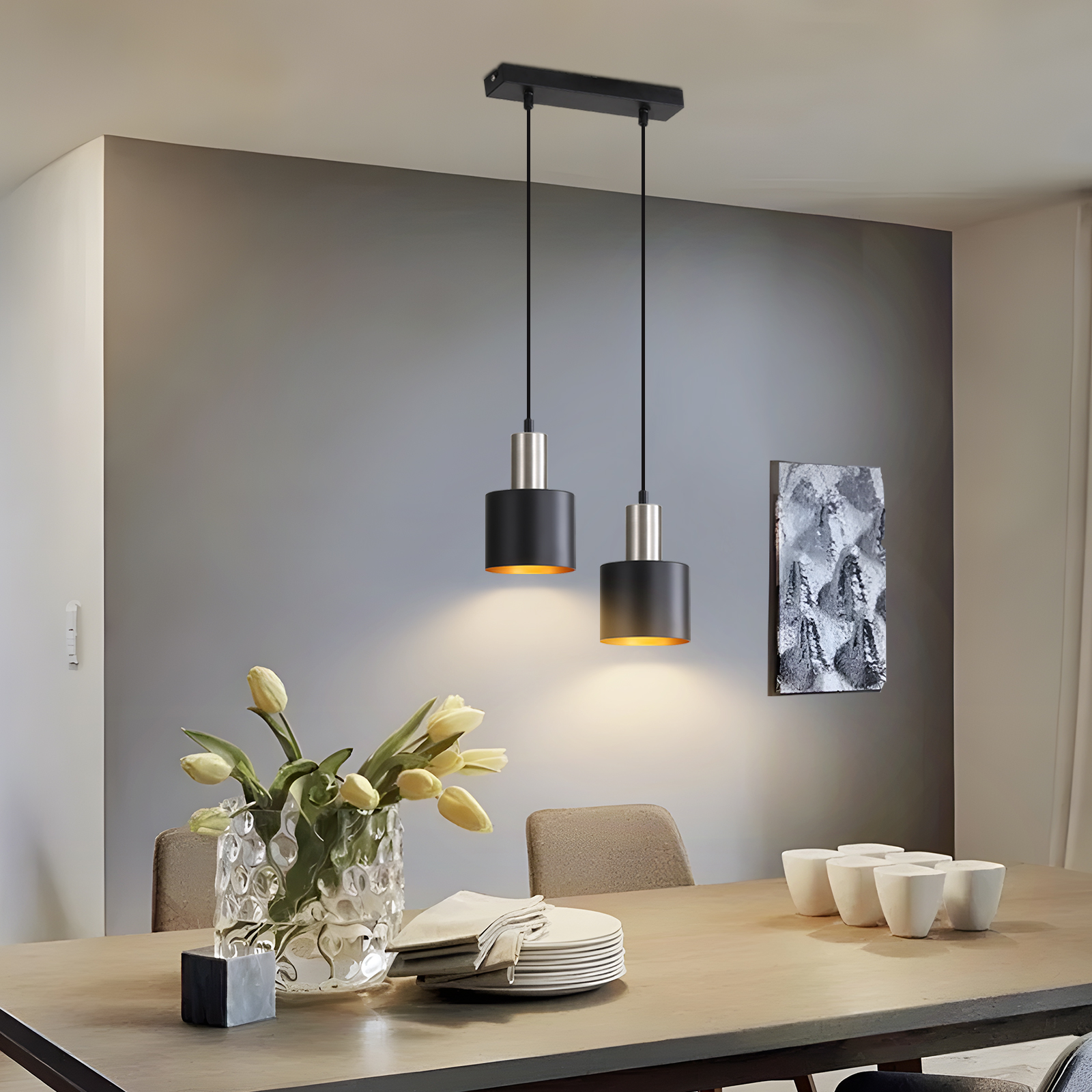 Esszimmer mit Tisch, zwei Hängelampen, Vase mit Blumen, Tellern, Tassen und einem Kunstwerk an der Wand.