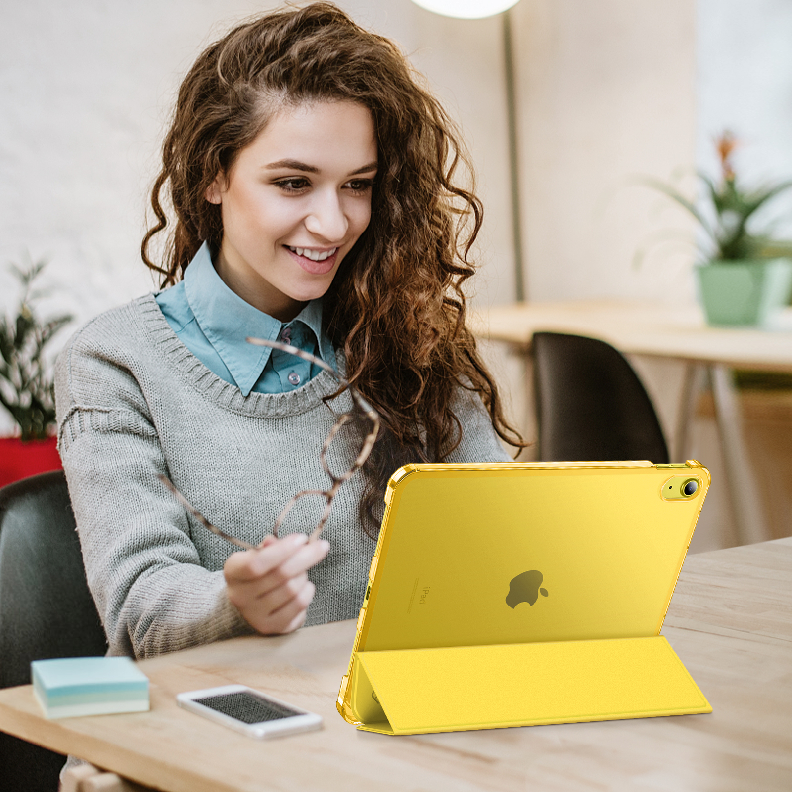 Frau lächelt, hält Brille, schaut auf gelbes iPad. Telefon und Haftnotizen auf dem Tisch.