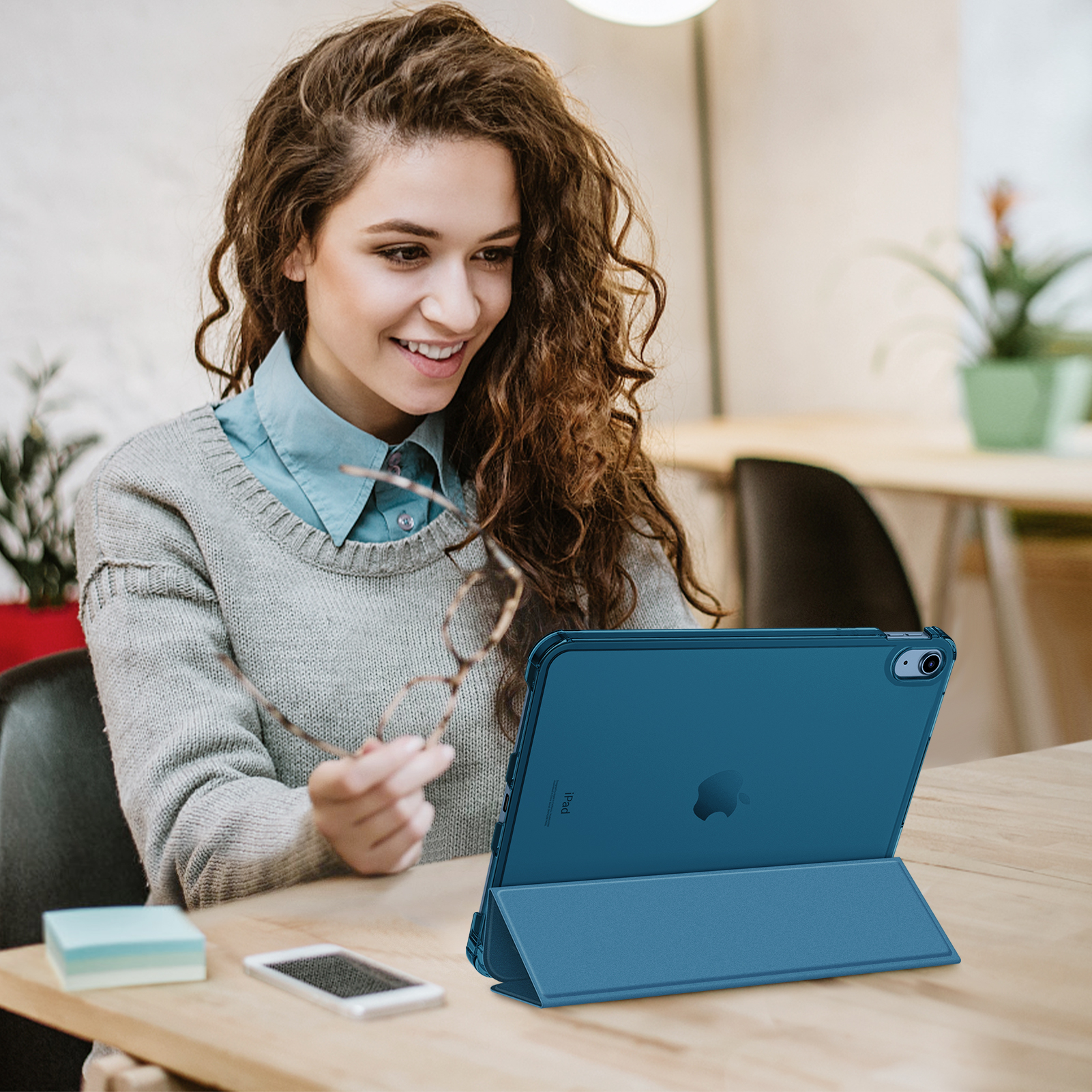 Eine Frau hält eine Brille und lächelt einen blauen Tablet-PC an. Ein Telefon und Haftnotizen liegen auf dem Schreibtisch.